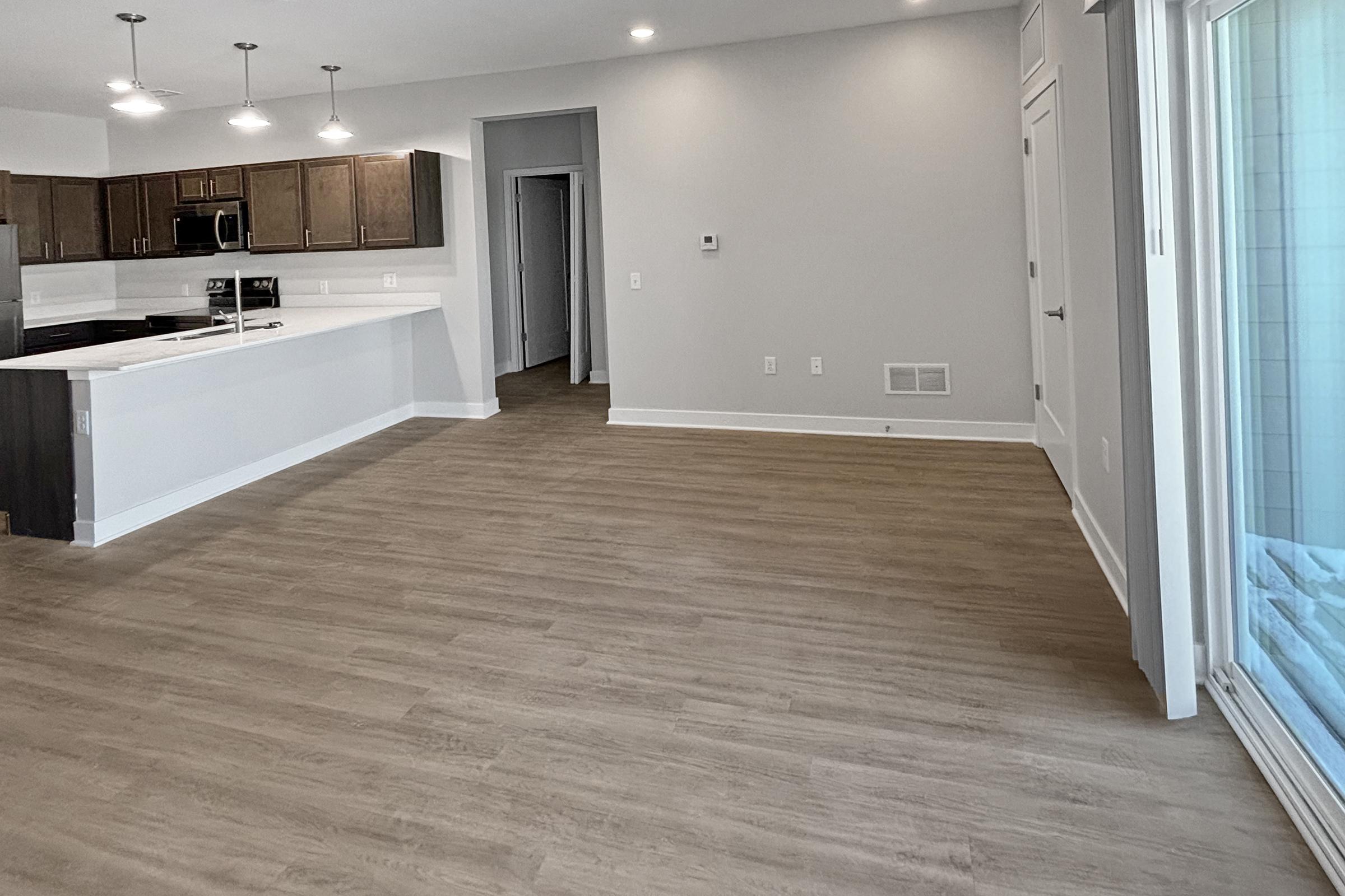 A spacious, modern living area featuring light-colored walls and wood-like flooring. The kitchen is visible with dark cabinetry and white countertops. Natural light enters through large windows, and there is an open layout leading to a hallway and an exit to a patio or balcony area.