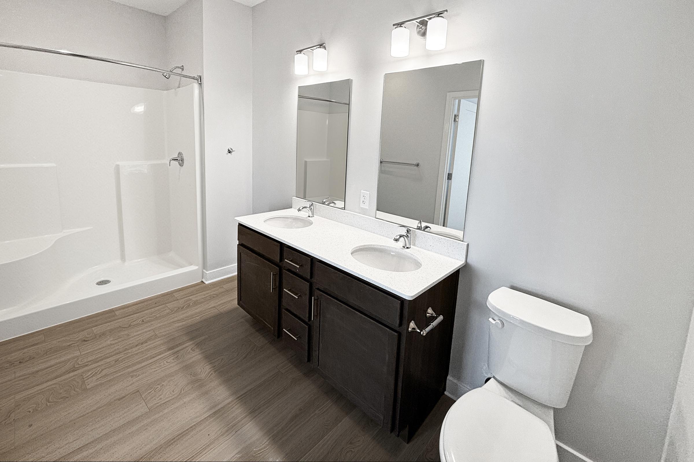 Modern bathroom featuring a double vanity with two mirrors, a white countertop, dark wooden cabinetry, and a toilet. The room has light grey walls, a large shower with a glass door, and light brown laminate flooring. Natural light enhances the space's bright, clean appearance.