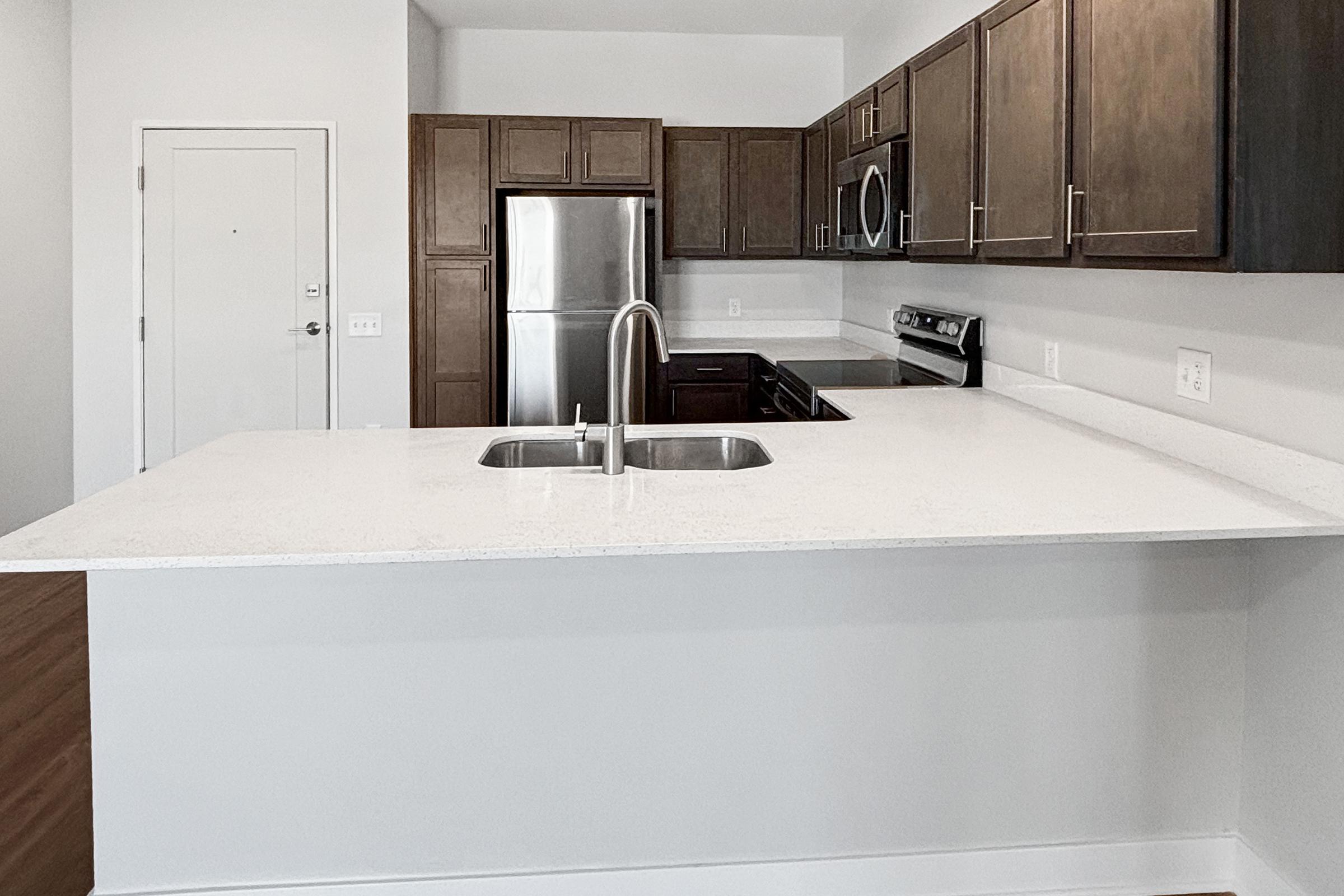 Modern kitchen featuring dark wooden cabinetry, stainless steel appliances, and a large island with a double sink. The open layout includes a refrigerator, stove, and microwave, with a clean, minimalist design. Light-colored countertops contrast with the dark cabinets, creating a bright and spacious feel.