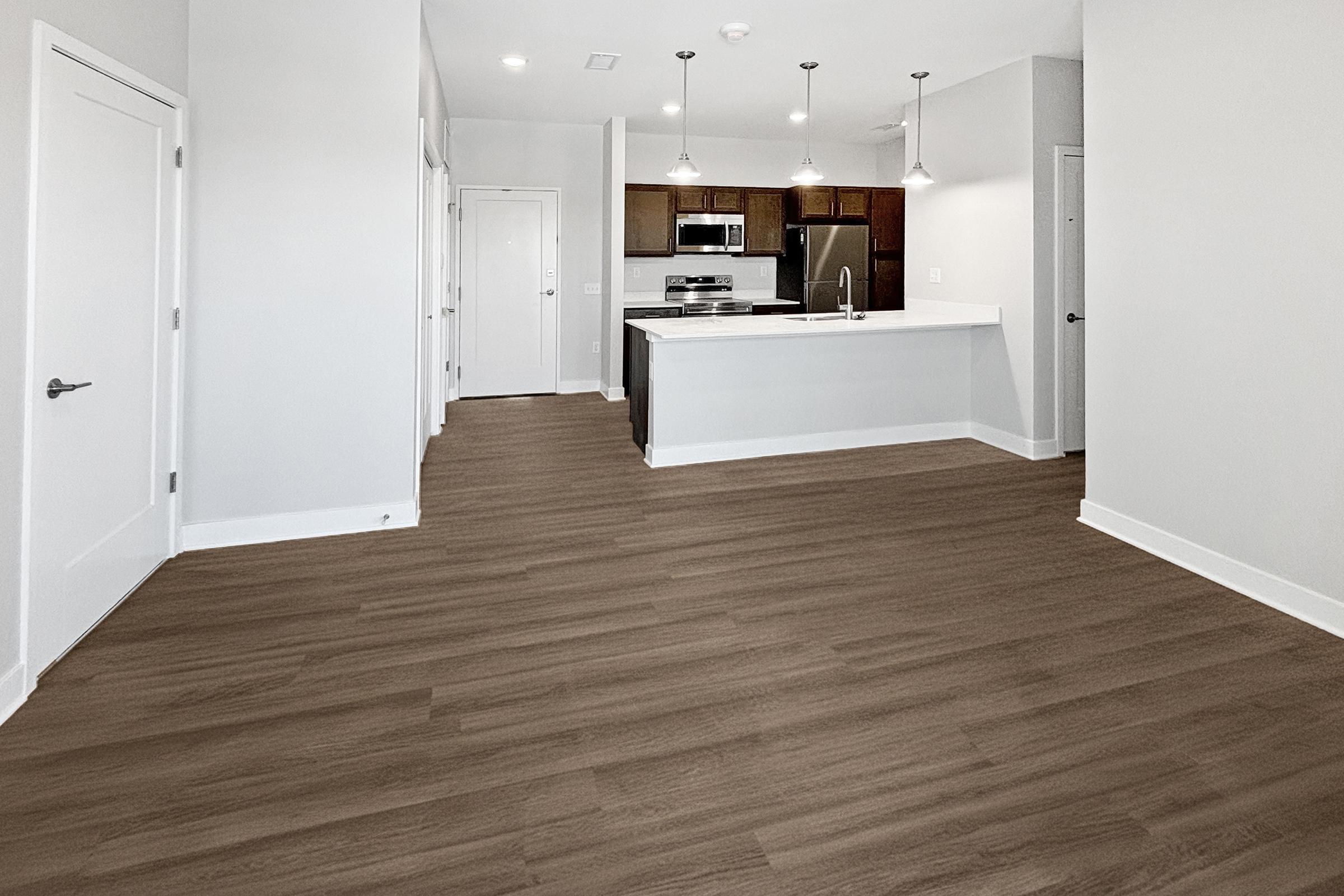 A spacious, modern apartment interior featuring a wooden-style floor, white walls, and a kitchen visible in the background with stainless steel appliances. The layout includes multiple doors and light fixtures hanging from the ceiling, creating a bright and inviting atmosphere.