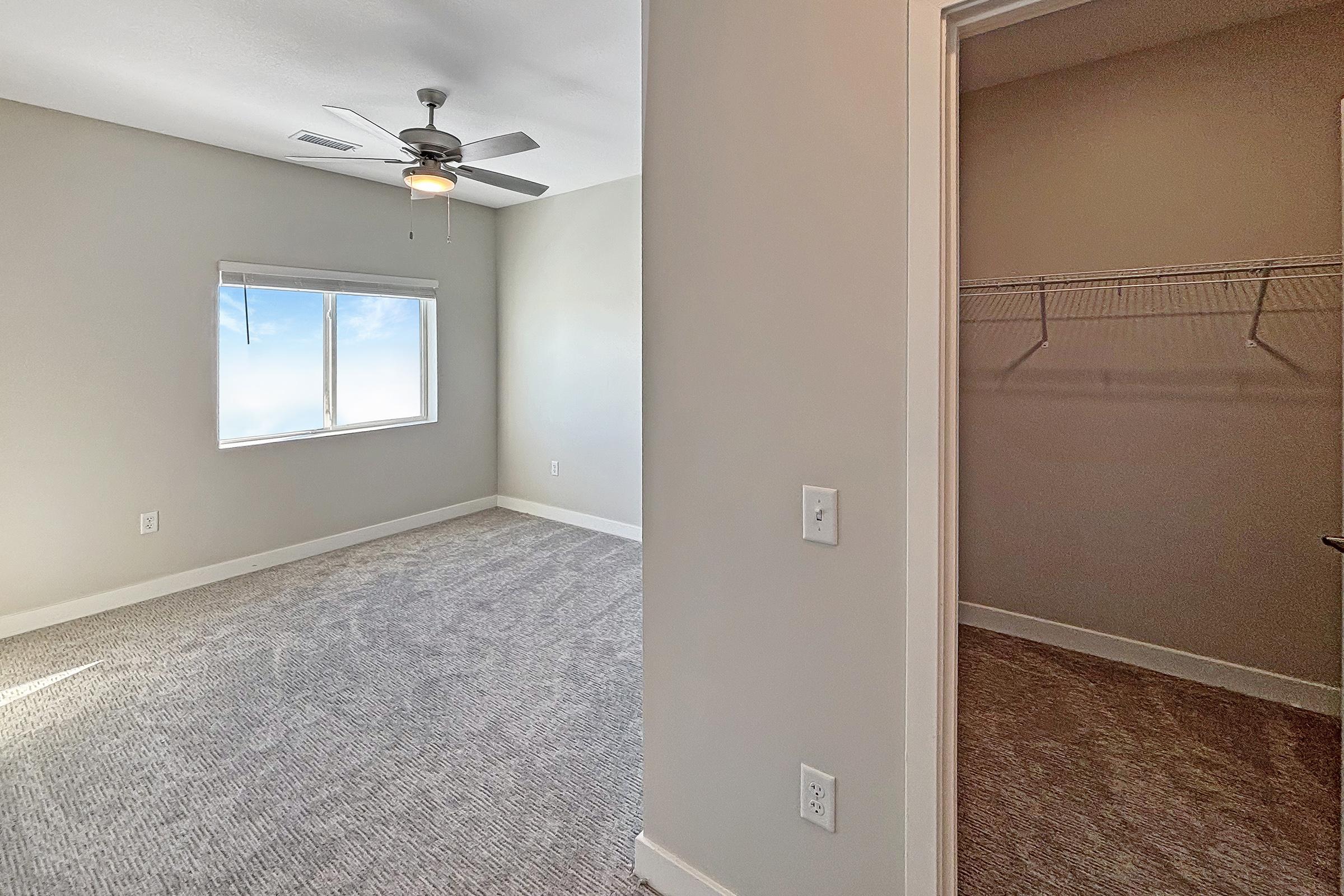 A bright room with light gray walls and carpet, featuring a ceiling fan and a large window that lets in natural light. To the right, an open closet with a wire shelving unit is visible. The overall aesthetic is modern and spacious, suitable for a bedroom or office space.