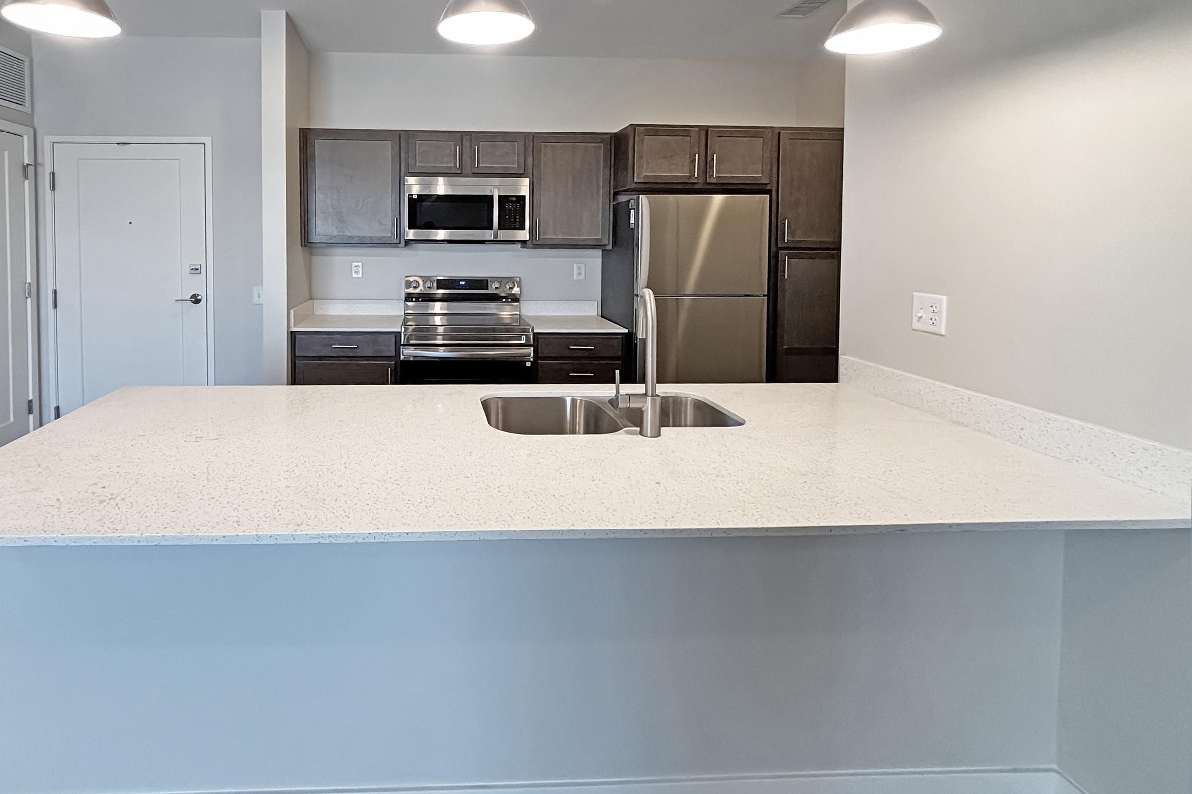 Modern kitchen featuring a large white countertop with a stainless steel sink, dark wood cabinets, and stainless steel appliances including an oven and refrigerator. Bright overhead lighting adds to the spacious and airy feel of the room.