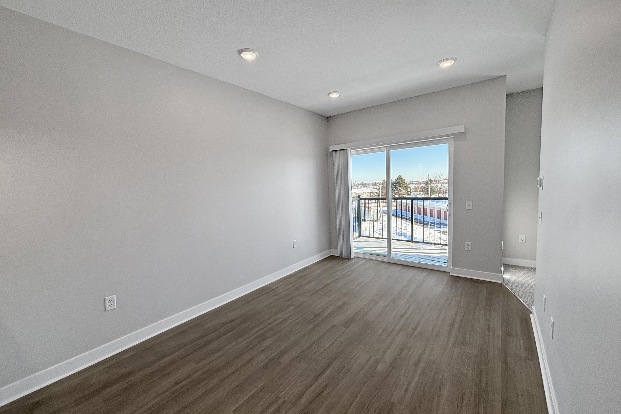 A bright, empty room with gray walls and wood-like flooring. It features a sliding glass door leading to a balcony with a view of the outdoors. Recessed lighting is installed in the ceiling, and the space appears ready for furniture. The overall atmosphere is clean and modern.