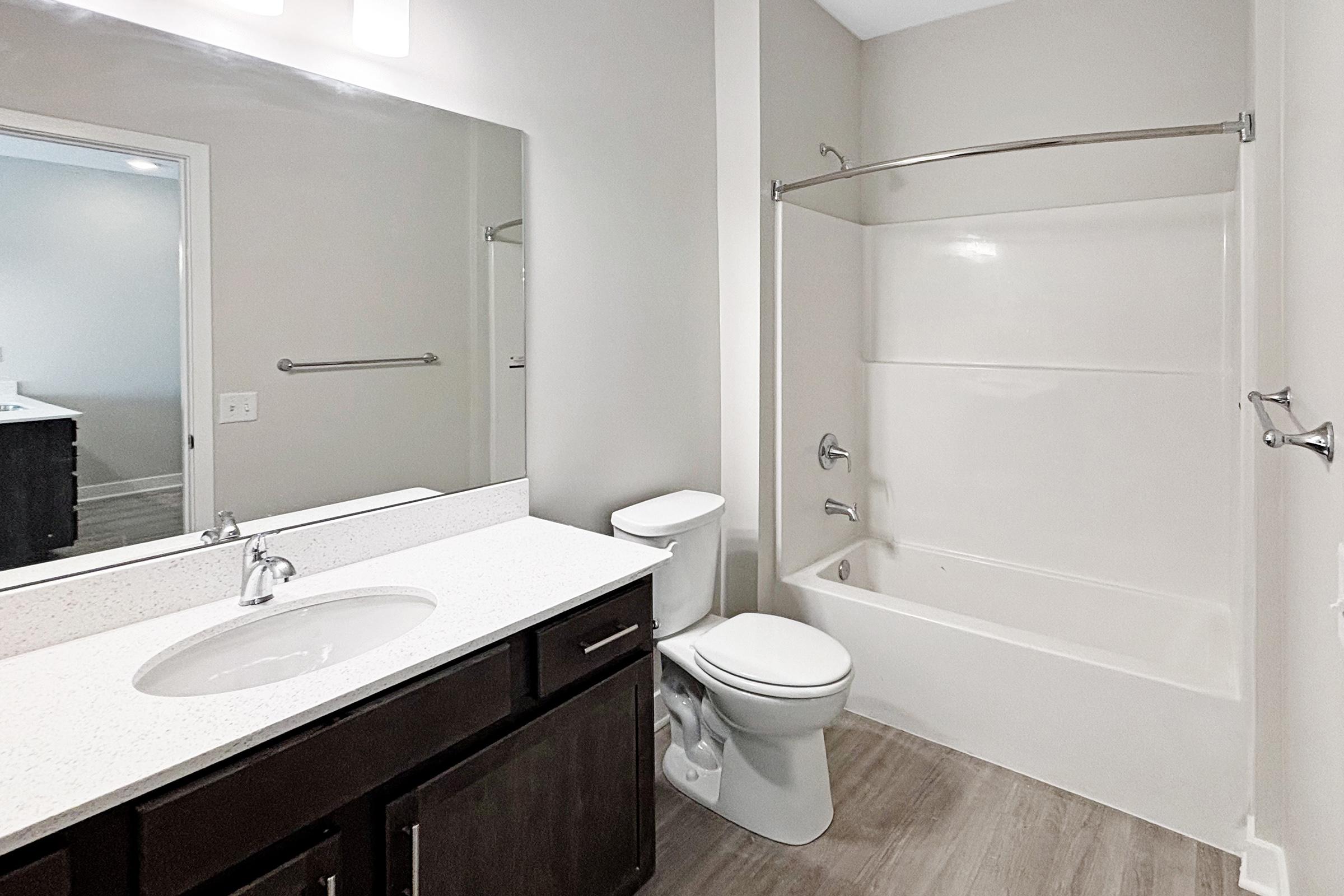 A modern bathroom featuring a large mirror above a vanity with a sink, dark wood cabinetry, a white bathtub with a shower curtain rod, and a toilet. The walls are painted in a neutral color, and the flooring is a light wood grain.