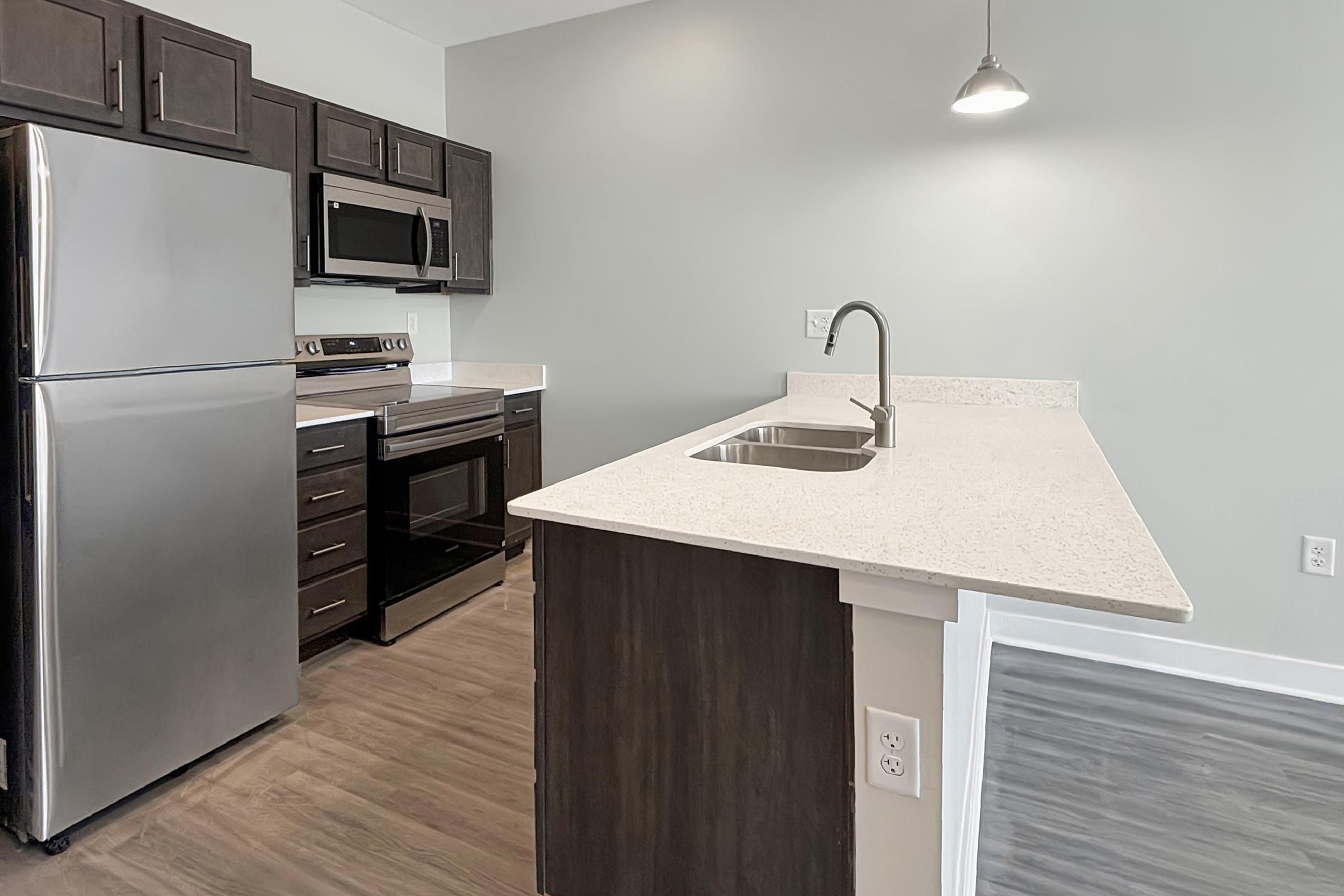 Modern kitchen featuring stainless steel appliances, including a refrigerator, oven, and microwave. The kitchen island has a double sink and a light-colored countertop. Dark wooden cabinets provide storage space, and the flooring is a light wood tone. The walls are painted a soft gray.
