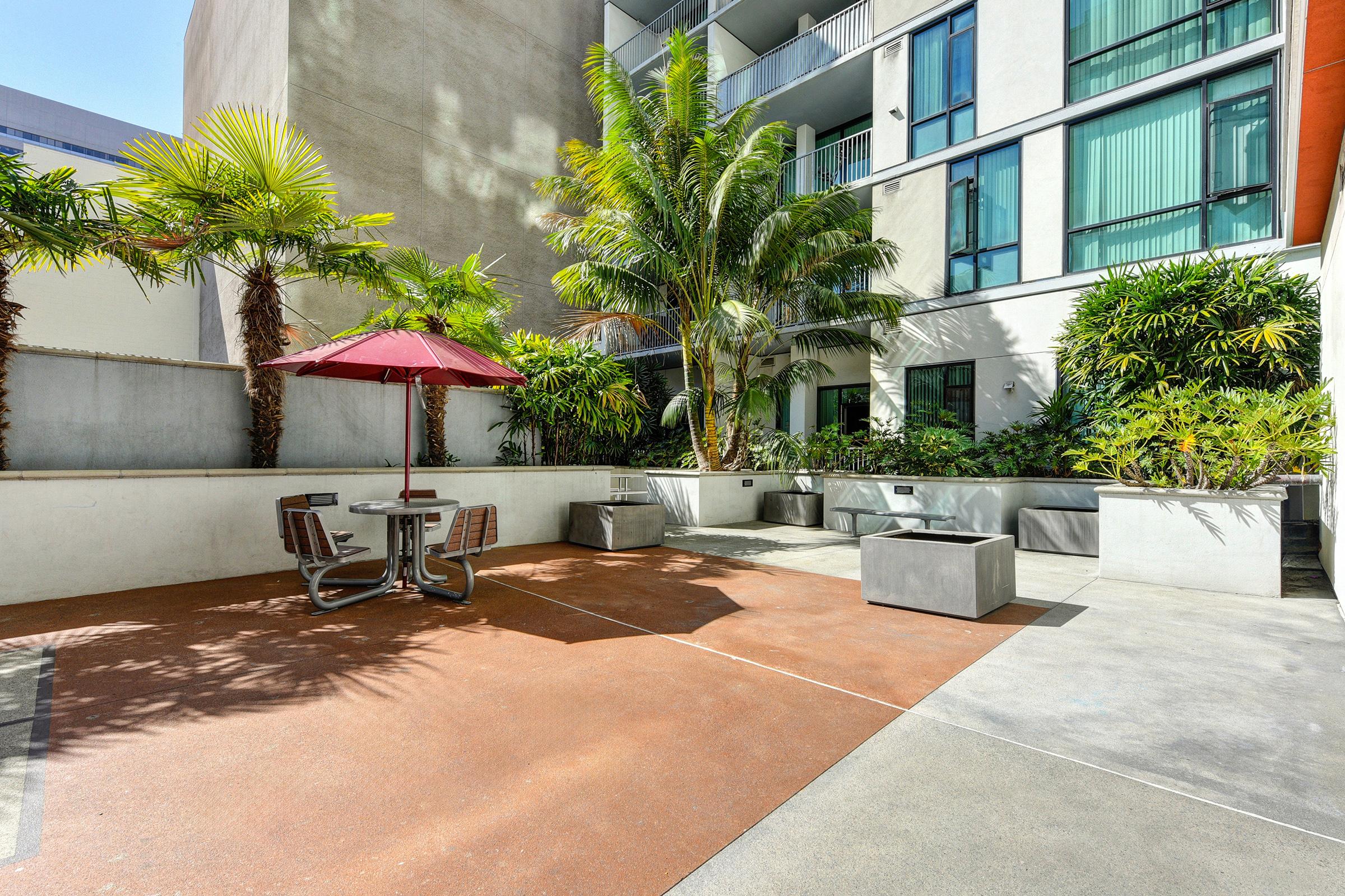Outdoor patio area featuring a reddish-brown surface, a table with seats, and a red umbrella for shade. Surrounded by lush greenery and palm trees, the space is part of a modern building with large windows reflecting light. Ideal for relaxation or social gatherings.