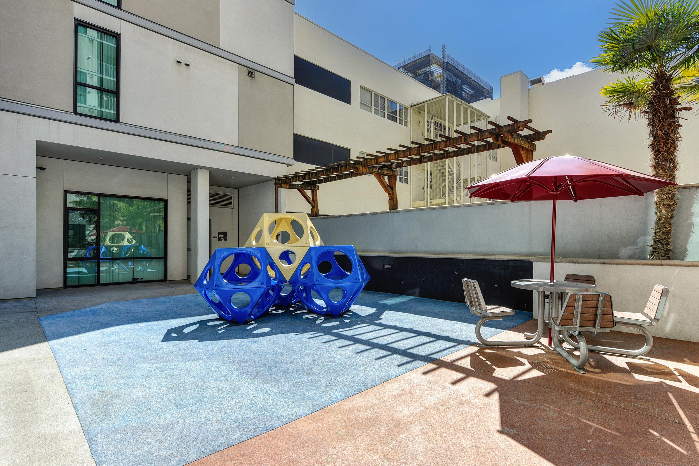 A vibrant outdoor play area featuring geometric climbing structures in blue and yellow, surrounded by textured flooring. A shaded picnic table and a red umbrella offer seating, while modern buildings and greenery add to the inviting atmosphere. Bright blue skies enhance the sunny, cheerful environment.