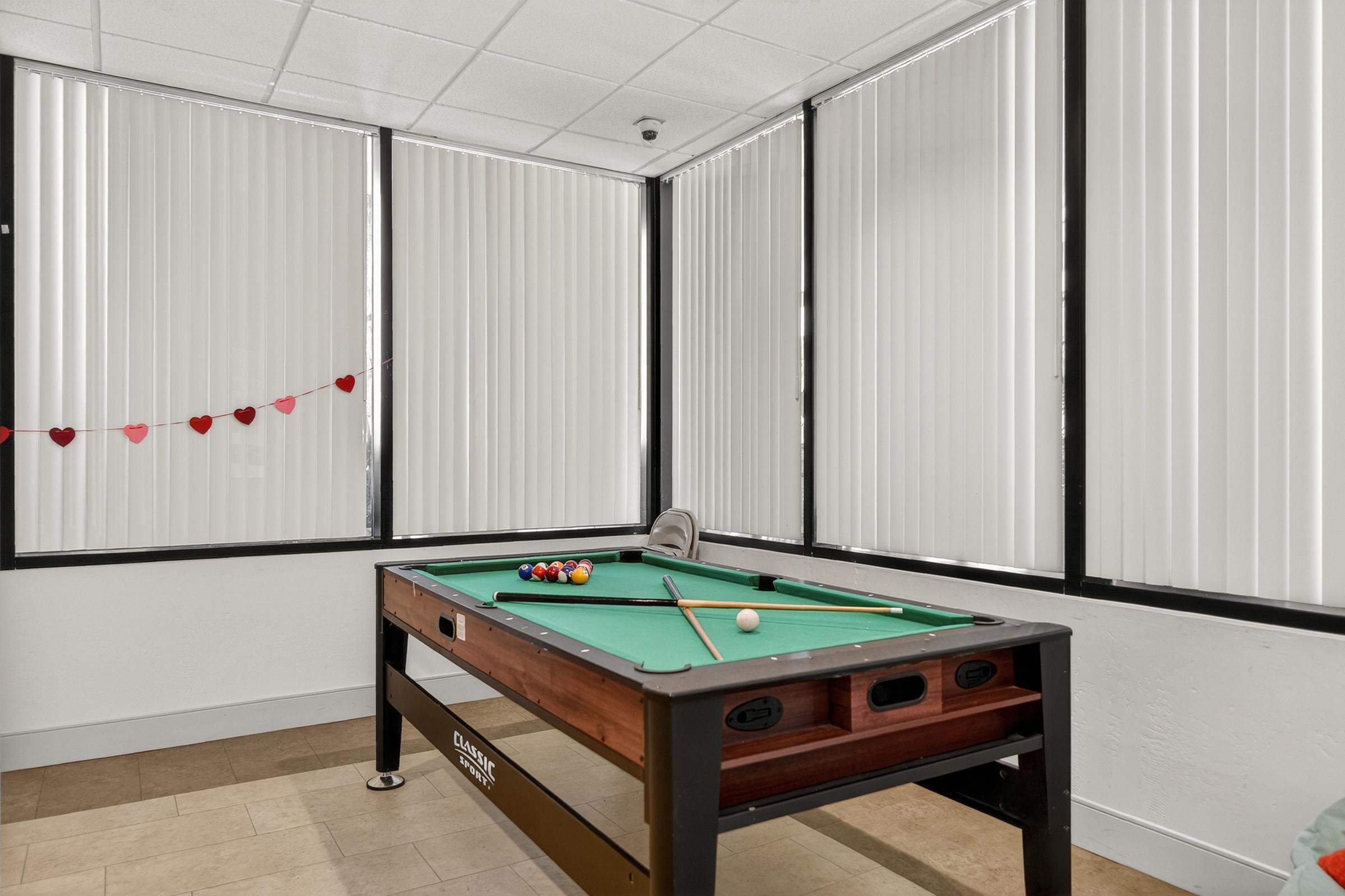 A pool table with a green felt surface, cue sticks, and several colorful billiard balls arranged on it. The table is placed in a well-lit room with large windows covered by vertical blinds. A string of small, red heart decorations hangs on the wall nearby.