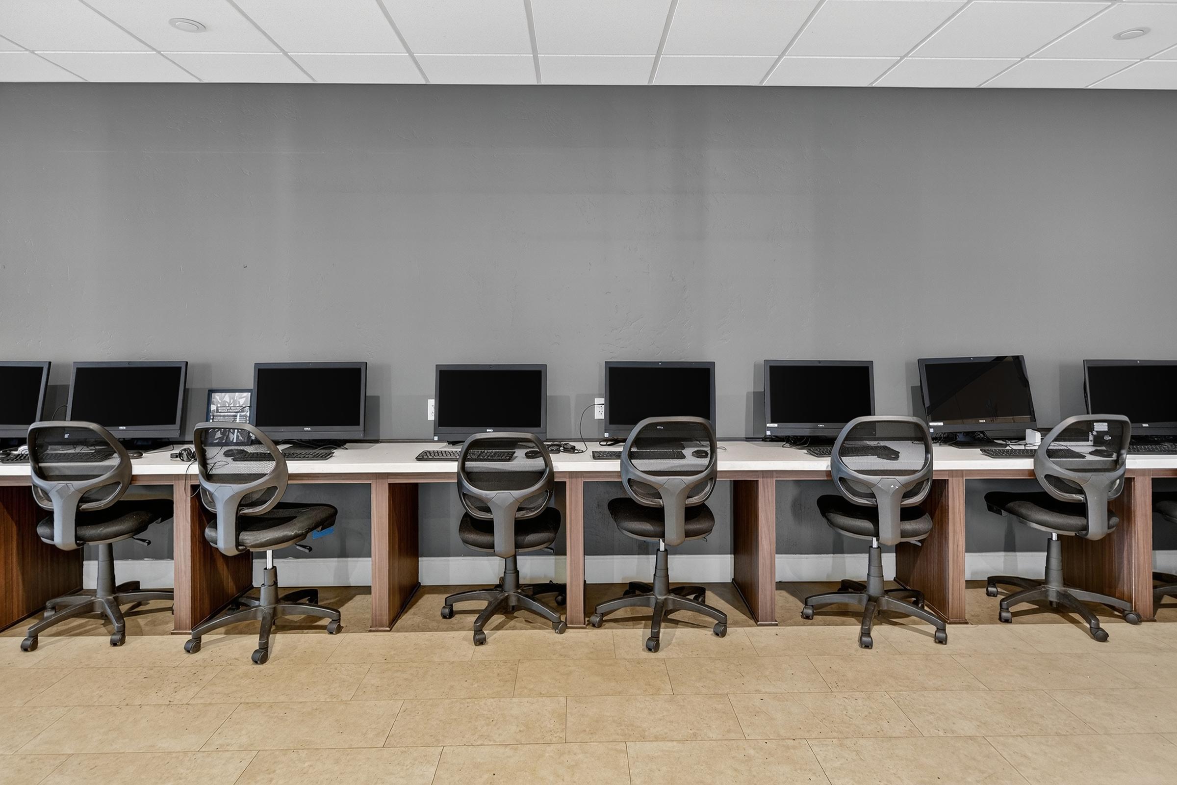 A row of empty computer workstations with black monitors, keyboards, and ergonomic chairs, set against a gray wall. The desks are wooden, and the space appears organized and ready for use. Natural lighting highlights the clean, modern design of the area.