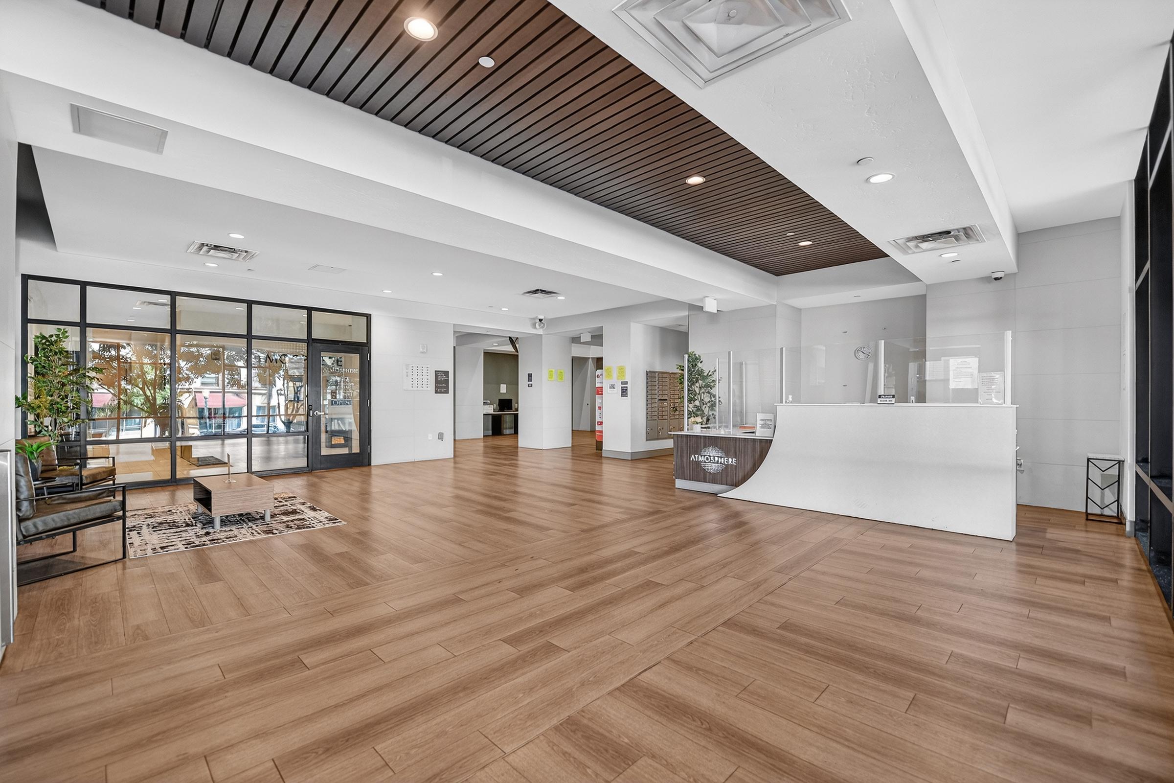 A modern reception area with wooden flooring and a white curved front desk. Large windows allow natural light to flood in, showcasing a spacious and inviting environment. Green plants add a touch of nature, and comfortable seating is arranged near the entrance. The overall design is sleek and contemporary.