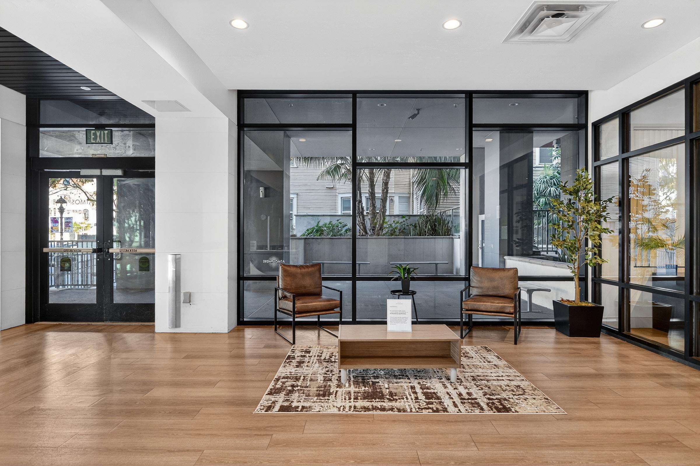 A modern lobby area featuring two brown chairs arranged around a low wooden coffee table on a patterned rug. Large windows allow natural light, showcasing greenery outside. The entrance is visible in the background, along with a stylish plant in a pot enhancing the ambiance.
