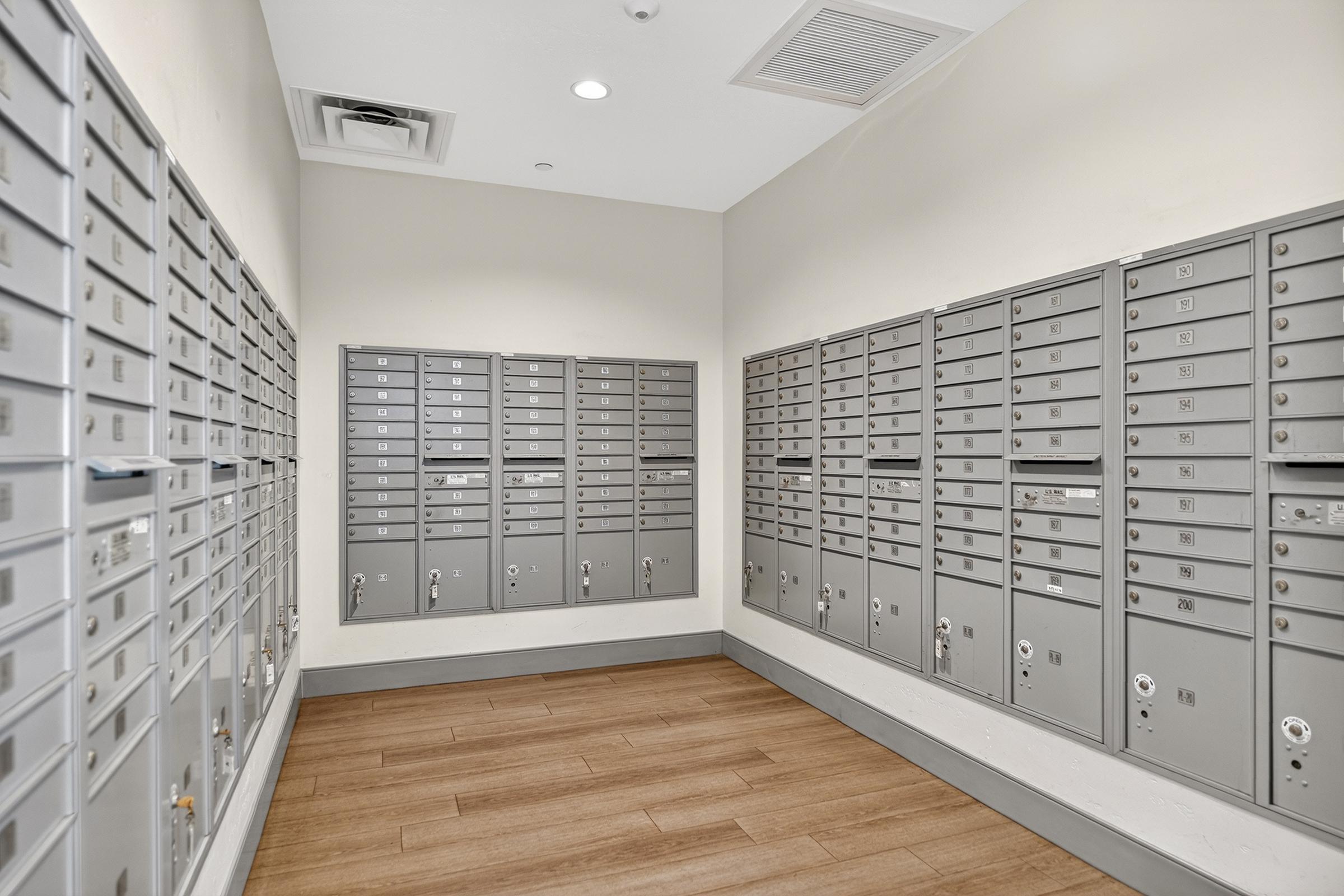 Interior view of a mailbox room featuring multiple gray mailboxes organized on two walls. The floor is made of wood, and the walls are painted in a light color. A ceiling panel and lighting fixtures are visible, creating a well-lit and functional space for residents to access their mail.