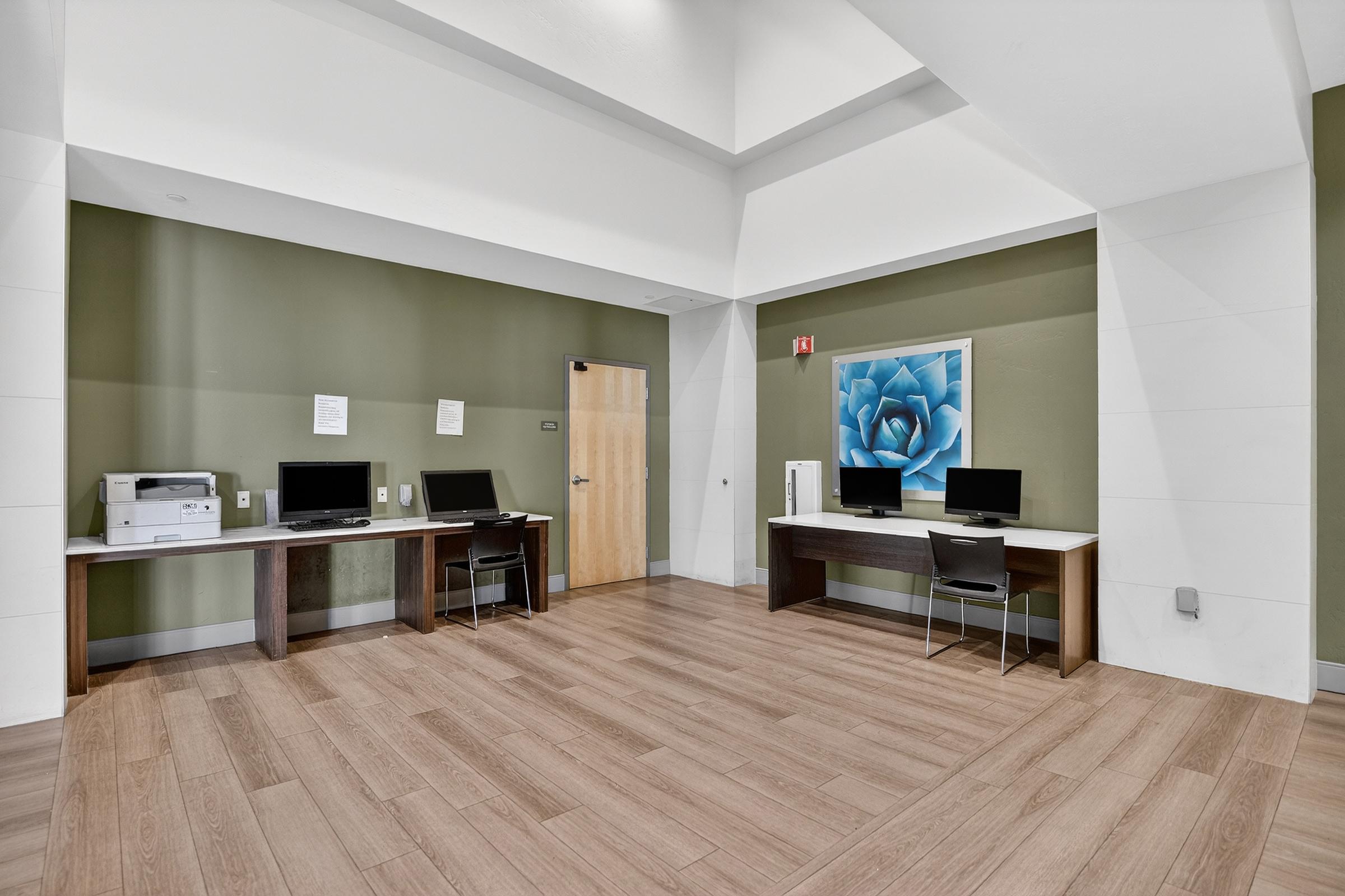 A well-lit study area featuring two desks with computer monitors, a printer on one desk, and a large wall art piece of a blue flower. The walls are painted in calming green, and the flooring is light wood. The space is modern and inviting, suitable for work or study.