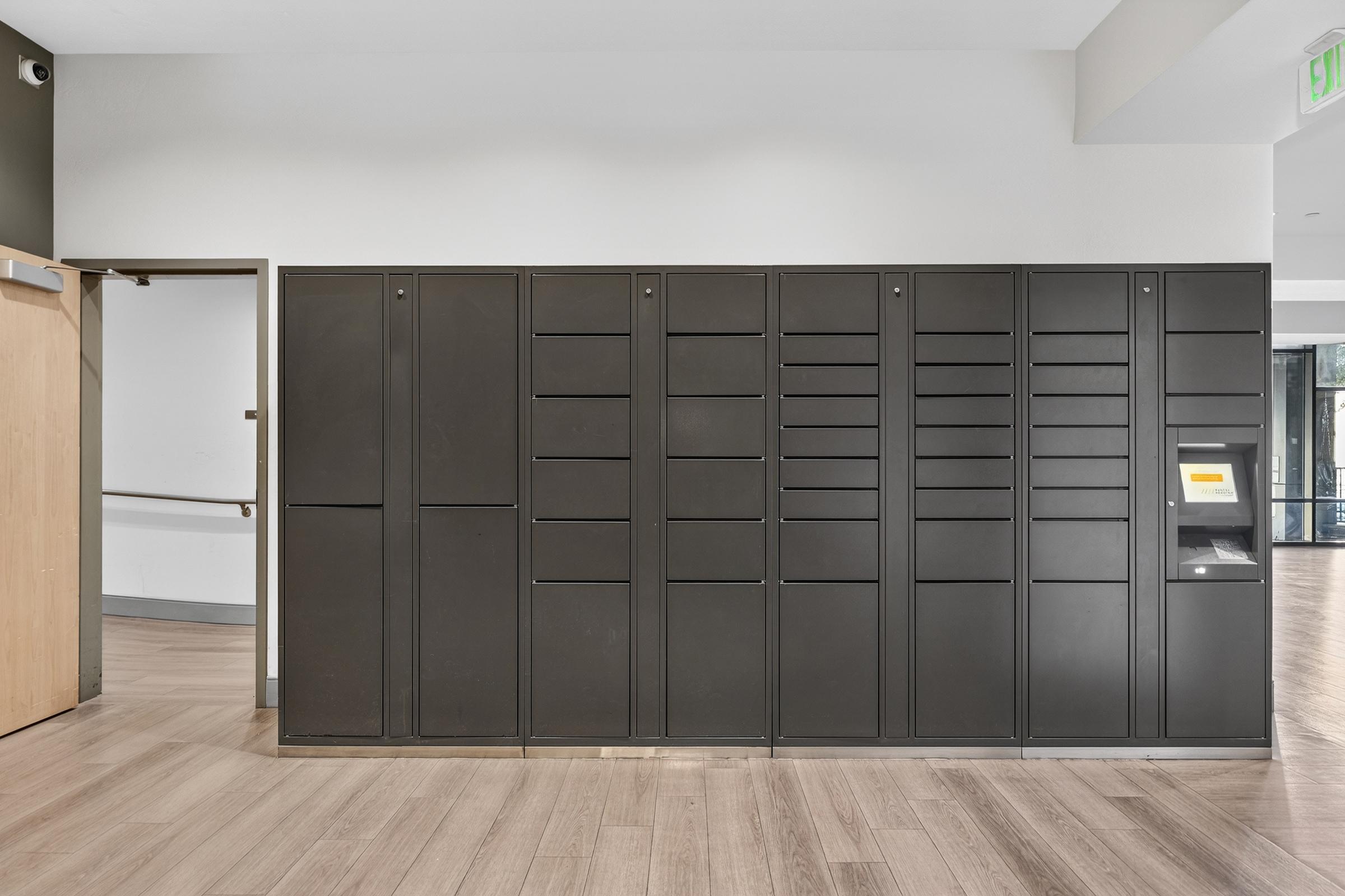 A row of black mail lockers mounted against a wall in a modern interior space. The lockers have various compartments, some of which are empty. On the right side, there is a small yellow and black kiosk for package retrieval. The flooring is light wood, and the overall ambiance is clean and contemporary.