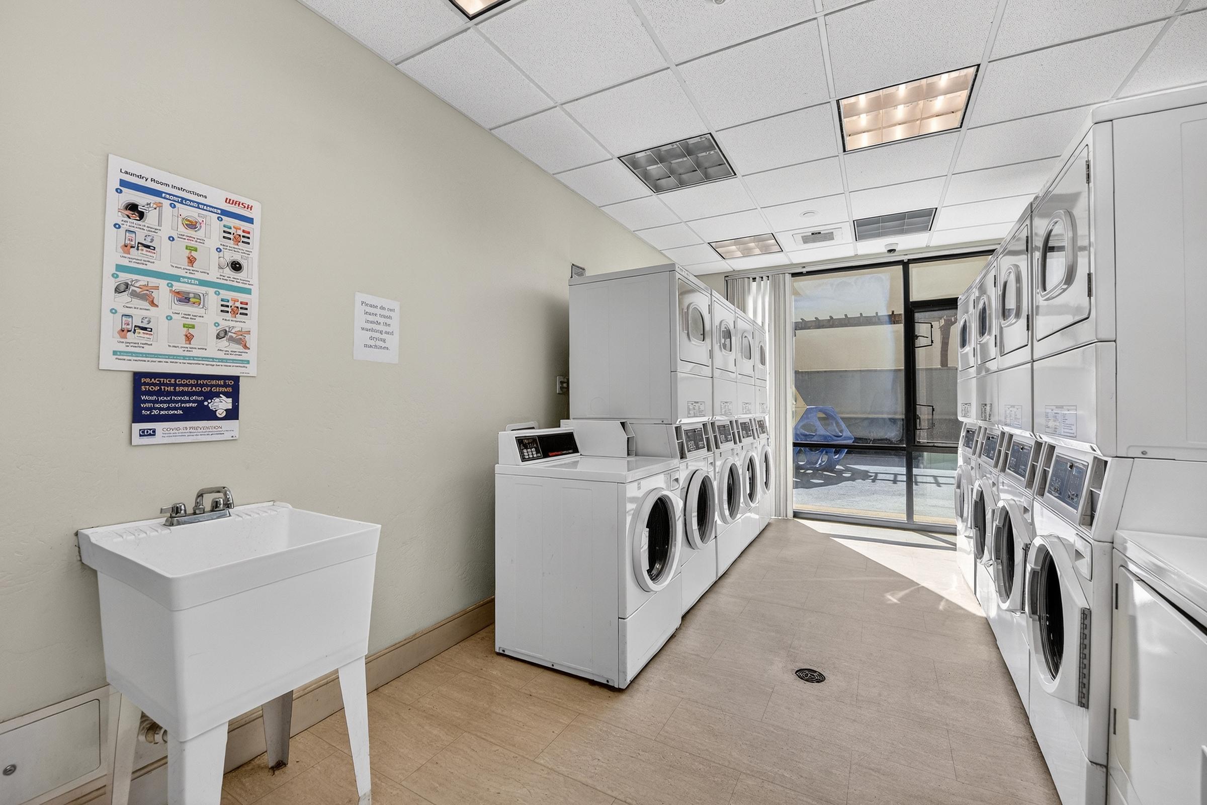 A clean and well-lit laundry room featuring a row of white washing machines and dryers along one wall. There is a utility sink on the left, and a set of instructions or guidelines is posted on the wall. Large windows provide natural light and a view of the outdoors.