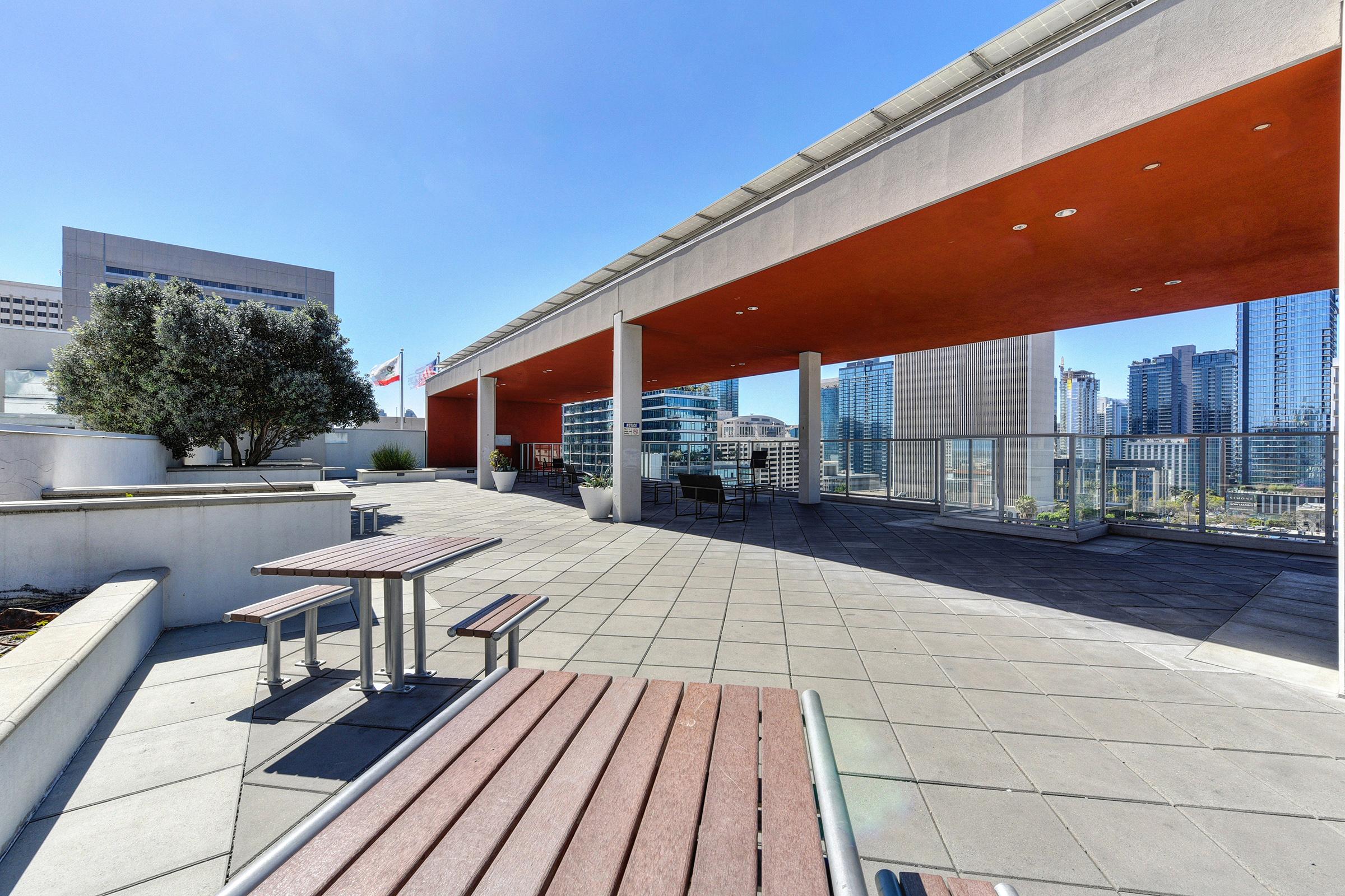 A modern outdoor rooftop space featuring a bright, sunny atmosphere. It includes wooden tables and benches, a sleek concrete surface, and green plants. The background showcases a city skyline with tall buildings and clear blue skies.