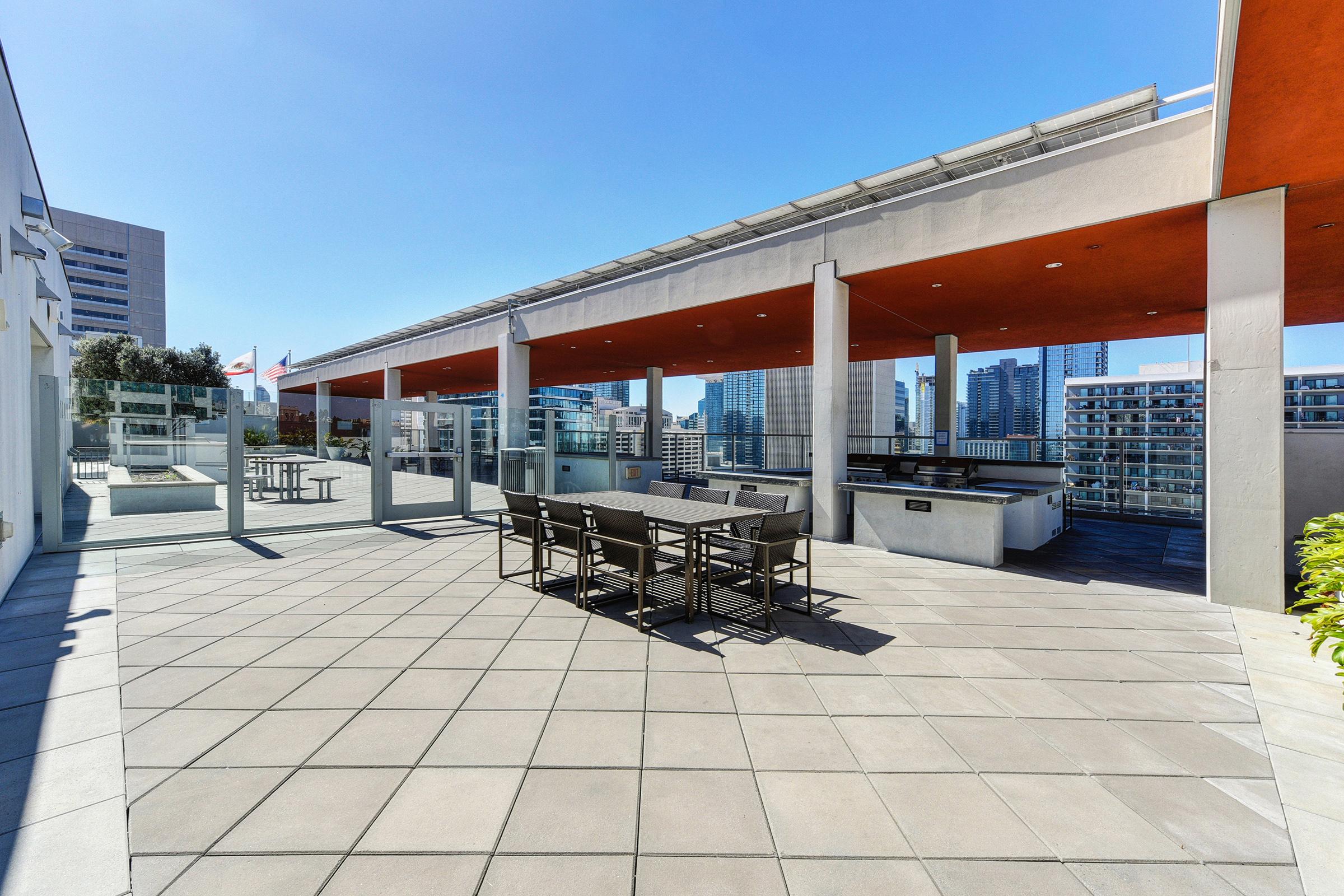 A modern rooftop terrace featuring a spacious patio with gray tile flooring, outdoor dining table, and barbecue area. The background showcases a city skyline under a clear blue sky, with buildings and greenery visible. Bright sunlight enhances the inviting atmosphere of the space.