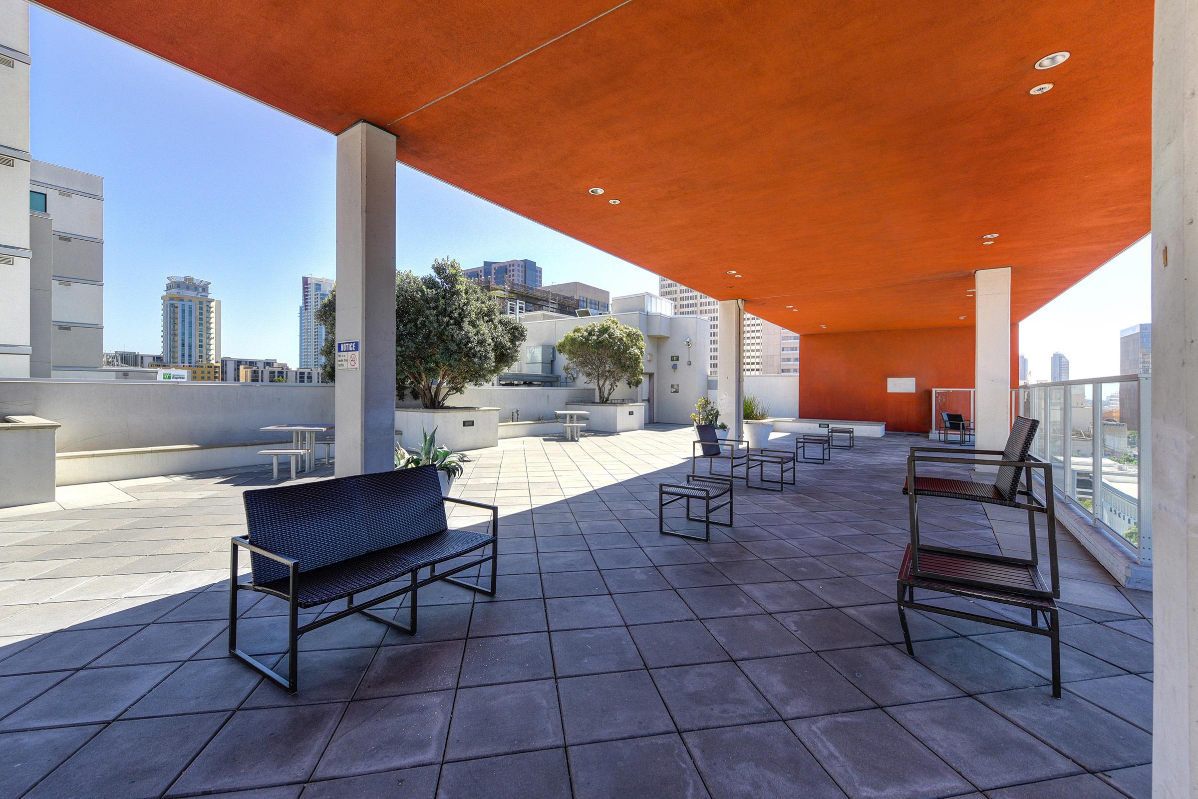A modern outdoor patio space with several black metal chairs and small tables. The floor is tiled, and there are planters with greenery. A vibrant orange ceiling contrasts with the sleek urban environment in the background, featuring tall buildings and a clear blue sky.