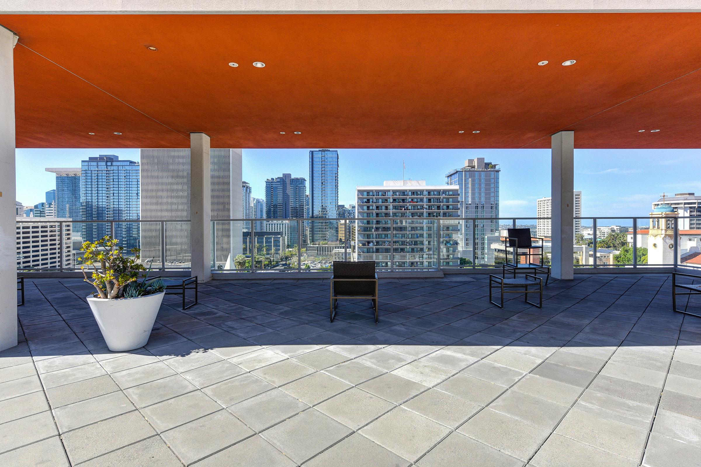 A rooftop terrace featuring modern chairs and a large potted plant, with a view of a city skyline. The area is shaded by an orange overhang, and the floor is tiled with geometric patterns. Skyscrapers and other buildings are visible in the background under a clear blue sky.