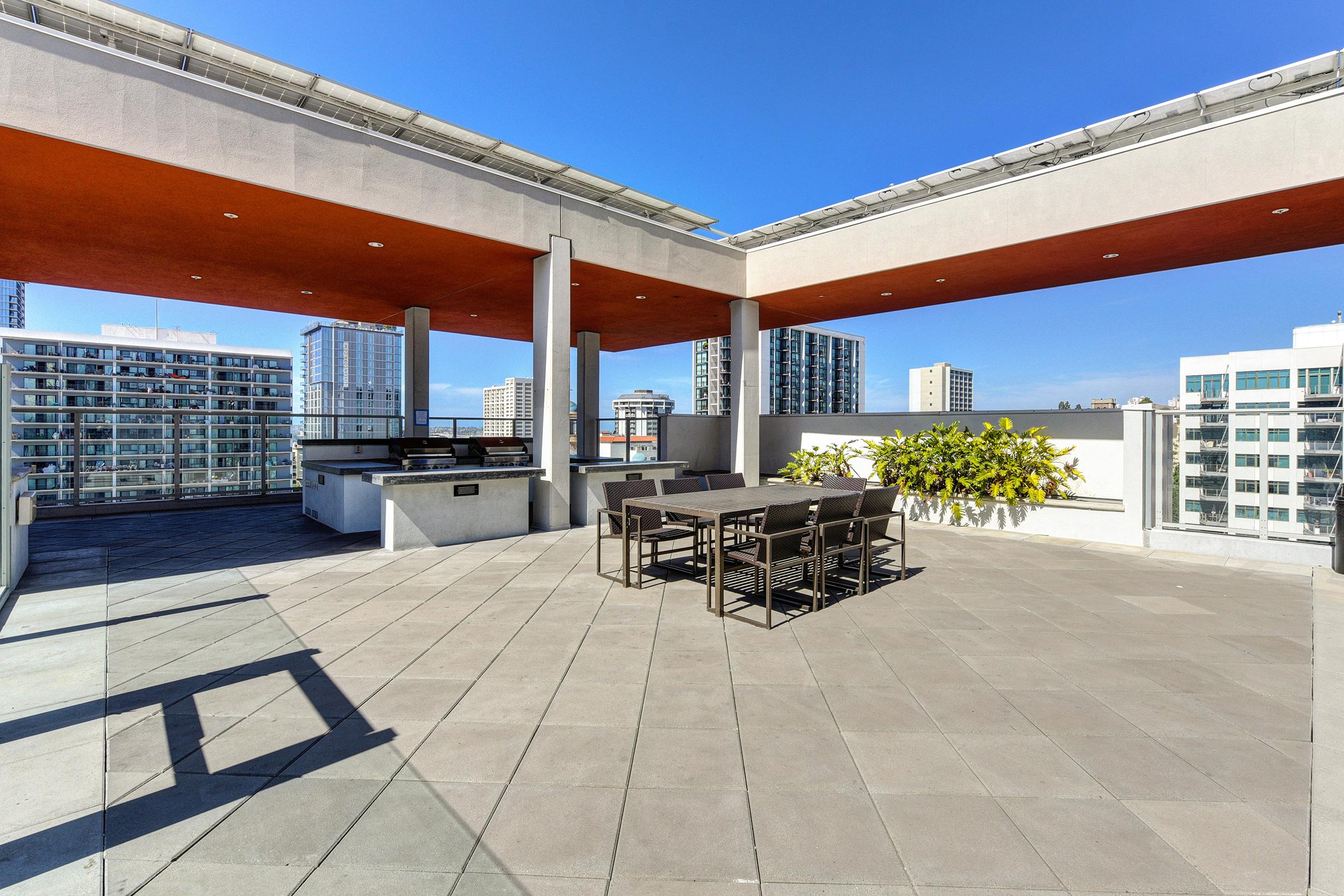 A modern rooftop terrace featuring a spacious layout with a dining table and chairs. Lush greenery in large planters adds a touch of nature. The surrounding skyline is visible in the background under a clear blue sky, creating a vibrant urban atmosphere.