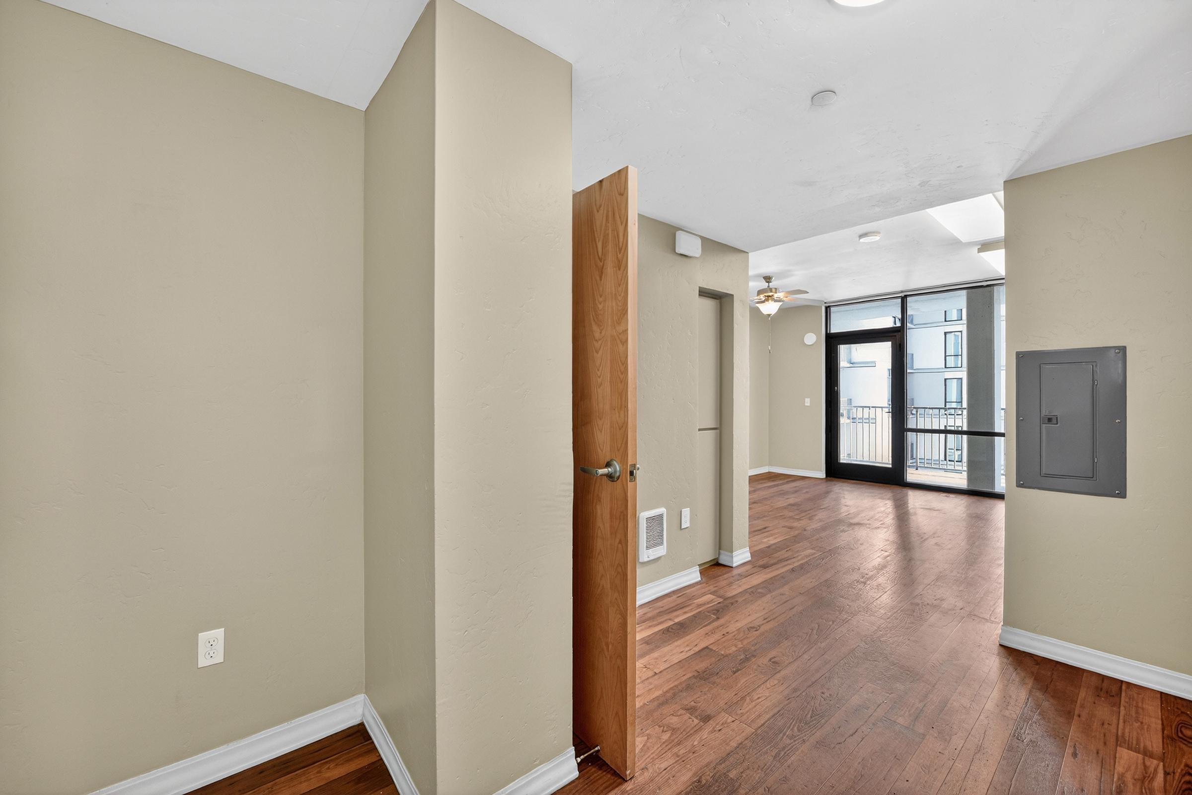A well-lit interior space featuring light beige walls, hardwood flooring, and a door partially open to another room. There are large windows with natural light, a ceiling fan, and a small utility panel on the wall. The layout appears open and modern, suggesting a living area or apartment.