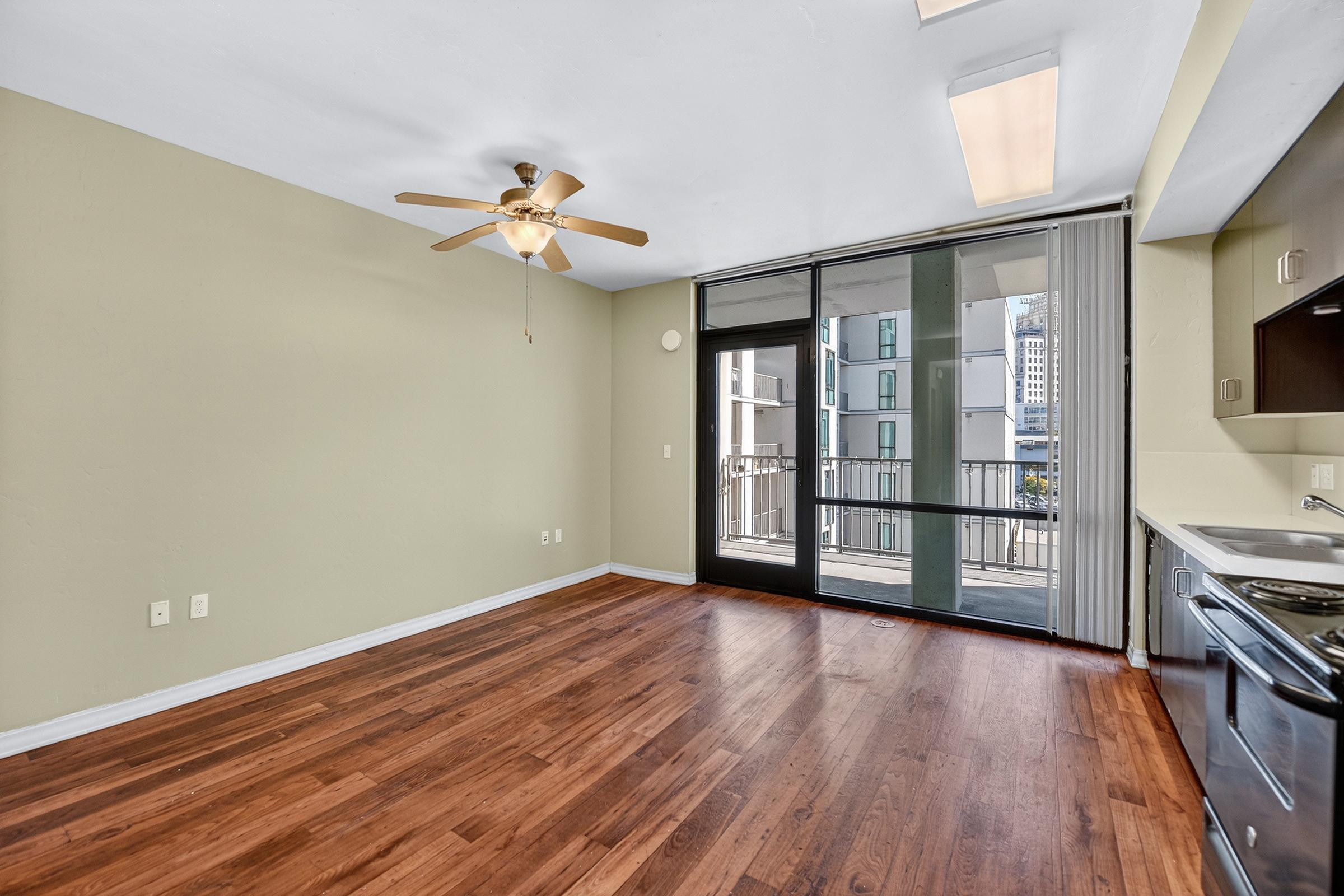 A spacious, well-lit living area featuring light green walls and hardwood floors. A ceiling fan is installed, and there are large sliding glass doors leading to a balcony with an urban view. The kitchen area is visible in the corner, showcasing modern appliances and cabinetry.