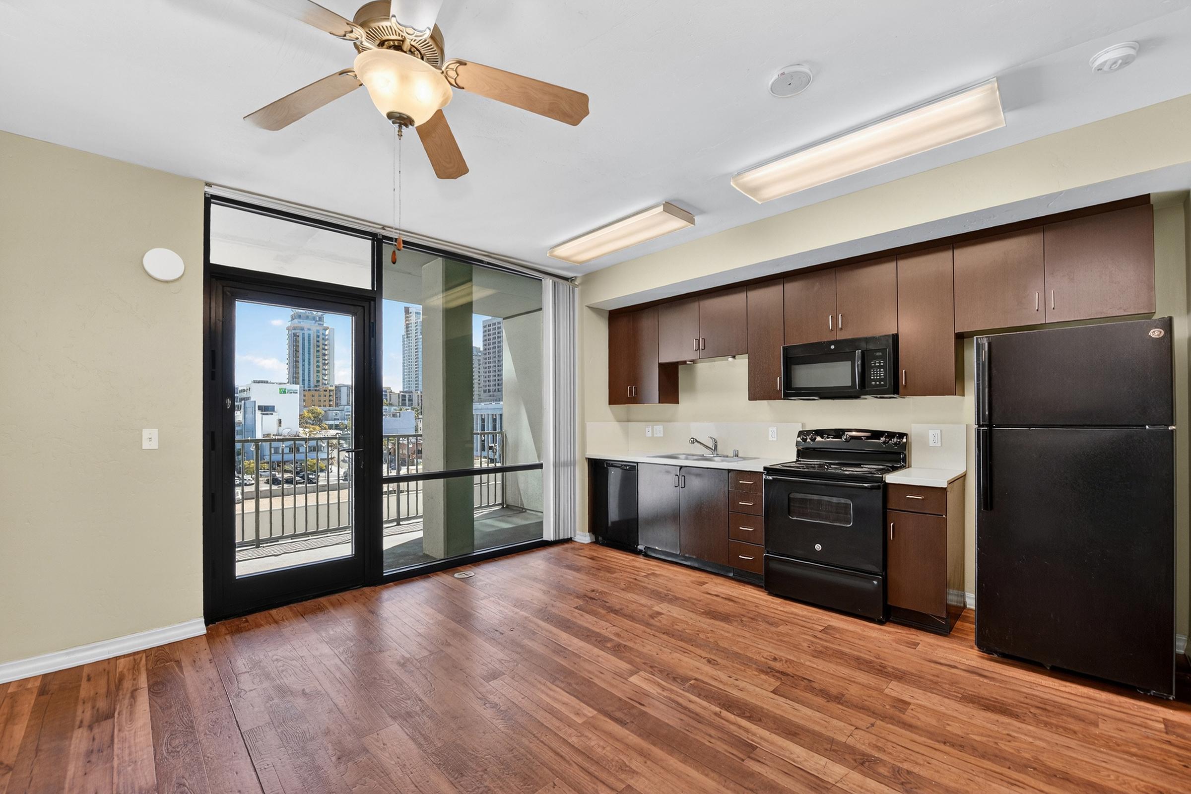 Bright kitchen and living area with wooden floors, featuring dark cabinetry, black appliances, and a ceiling fan. Large windows offer views of city buildings. The space has modern lighting and a sliding glass door leading to a balcony.