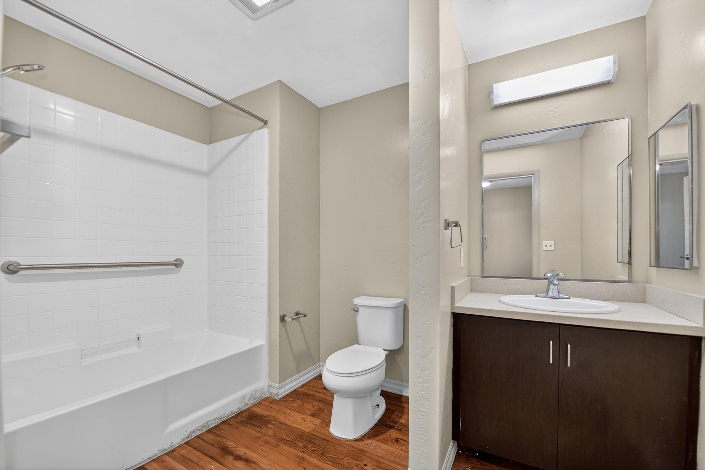 A clean, modern bathroom featuring a white bathtub with a showerhead, a toilet, and a single sink vanity with a silver faucet. The walls are painted in a neutral color, complemented by wood-style flooring. A large mirror is above the sink, with additional lighting above it.