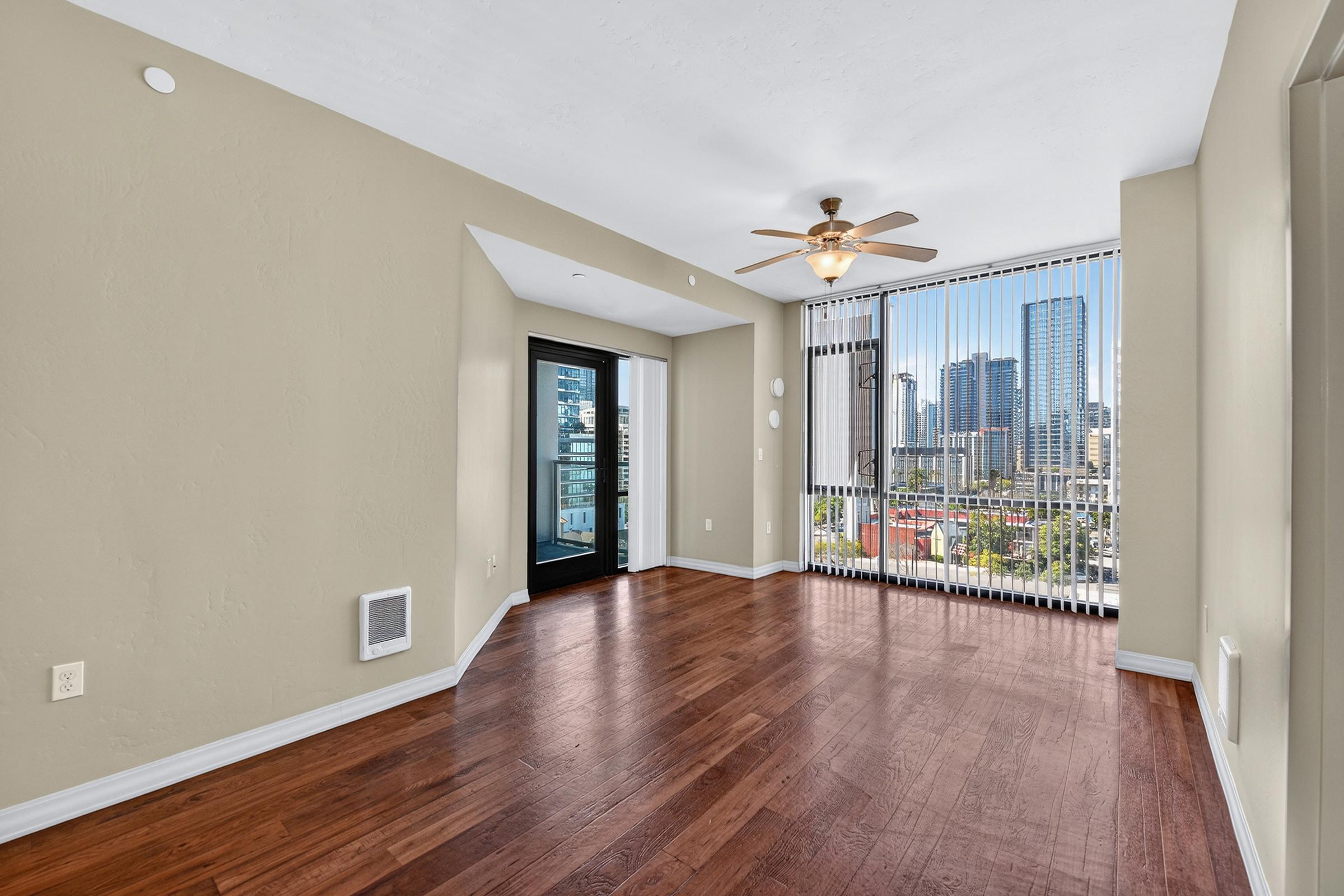 A spacious, well-lit living room featuring hardwood floors, light beige walls, and a ceiling fan. Large windows with blinds offer a view of a city skyline. A sliding glass door leads to a balcony. The room is empty, creating a clean, inviting atmosphere.