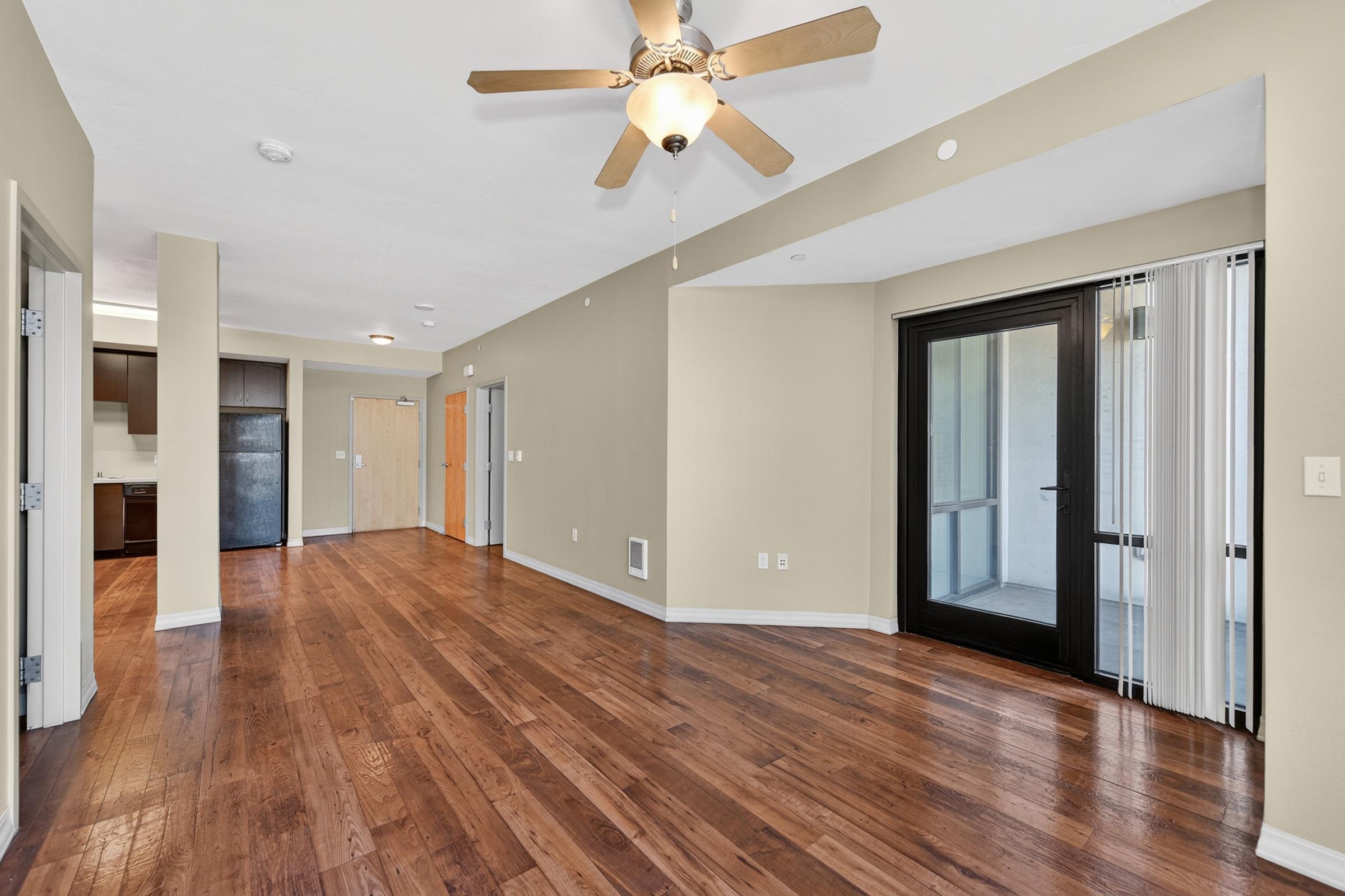 A spacious living area featuring polished hardwood floors, a ceiling fan, and a large glass door leading to a balcony or patio. The space is painted in neutral tones and includes a view into a kitchen area with dark cabinetry. Natural light enters through the glass door and windows.