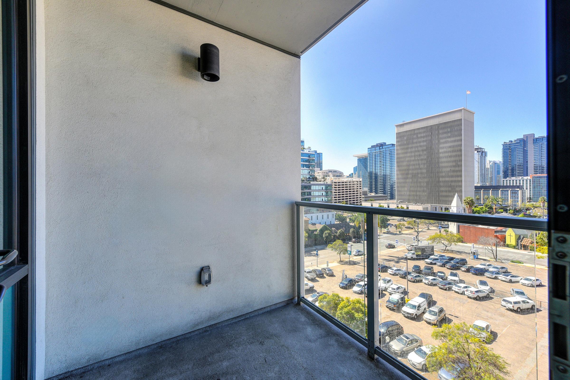 A view from a balcony looking out onto a cityscape, with modern buildings in the background, a clear blue sky, and a parking lot filled with cars below. The balcony has a glass railing and a light fixture mounted on the wall.