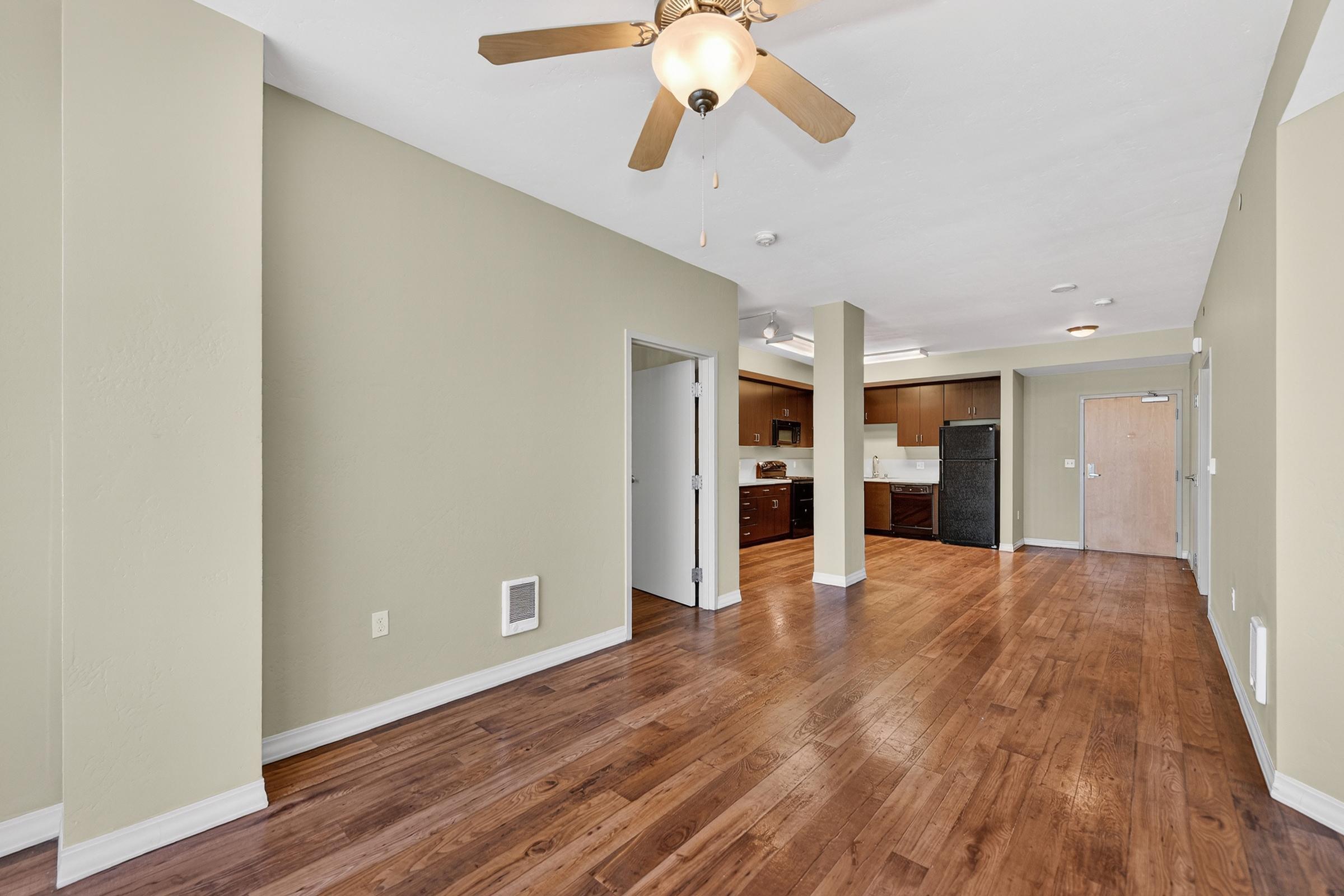 Spacious living area with hardwood floors, featuring a ceiling fan and light fixture. Open layout connects to a kitchen with dark wood cabinets and appliances. Light beige walls create a warm atmosphere. Doorway leads to a separate room, enhancing the openness of the space.