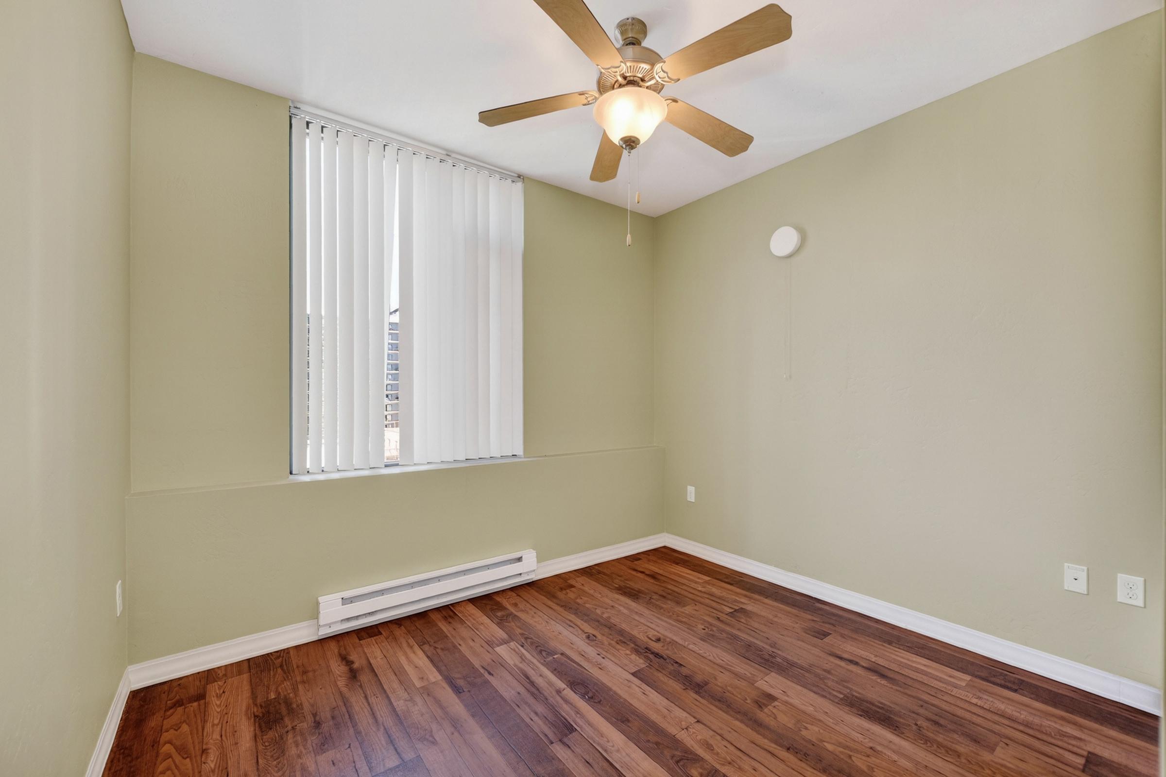 A vacant room with light green walls, hardwood flooring, and a ceiling fan. There's a window covered with vertical blinds allowing natural light. A wall-mounted heater is visible, and a small circular object is on the wall. The room has a minimalist design, making it suitable for various uses.
