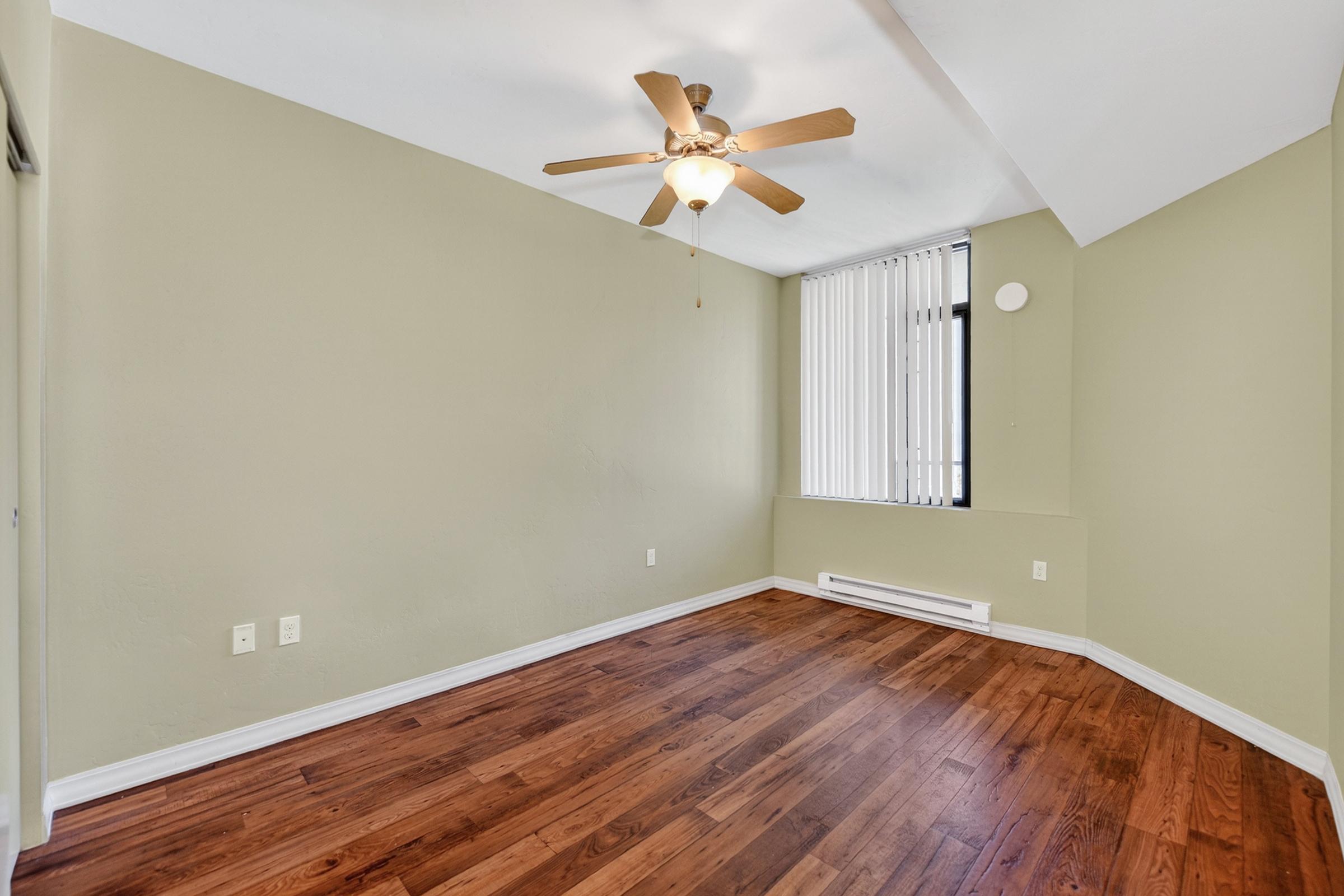 Empty room with light green walls, a ceiling fan, and hardwood flooring. A window with blinds is on one wall, and there’s a baseboard heater nearby. The room has a simple and spacious layout, making it suitable for various uses.