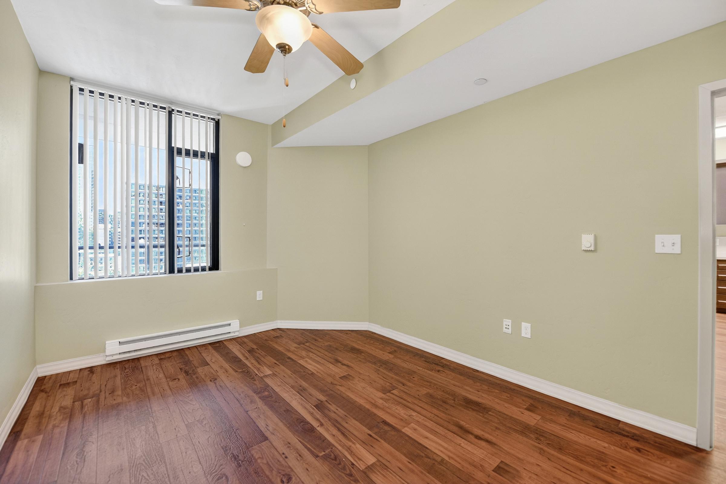 A well-lit room with a ceiling fan, featuring warm beige walls and wooden flooring. Large windows with vertical blinds allow natural light, while a small heating unit is visible on the wall. The room has a modern and spacious feel, suitable for various uses such as a living area or office.