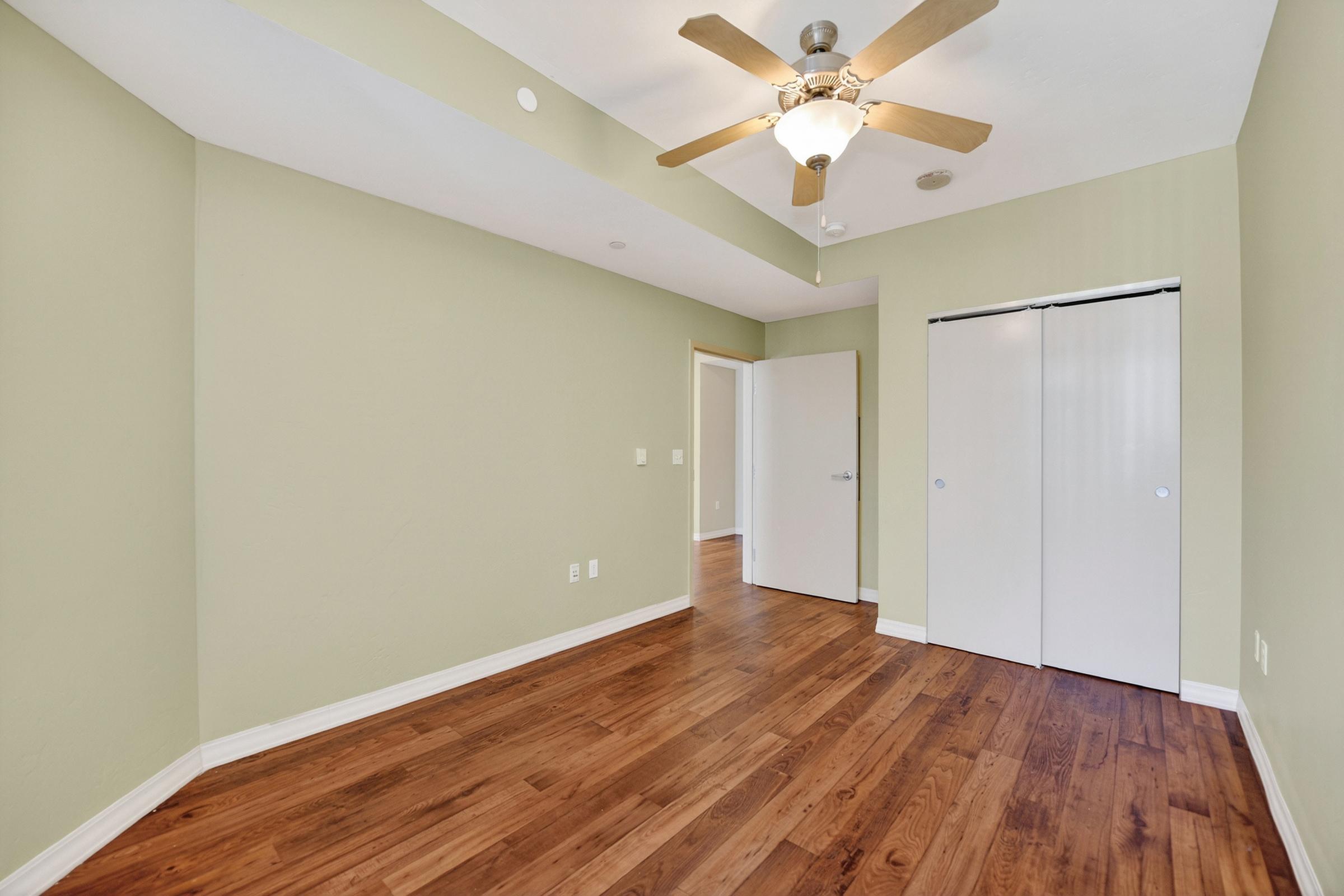 A spacious empty room with light green walls and wooden floors. It features a ceiling fan, a closed door leading to another space, and a two-door closet. Natural light enters through the doorway, creating a bright and inviting atmosphere.