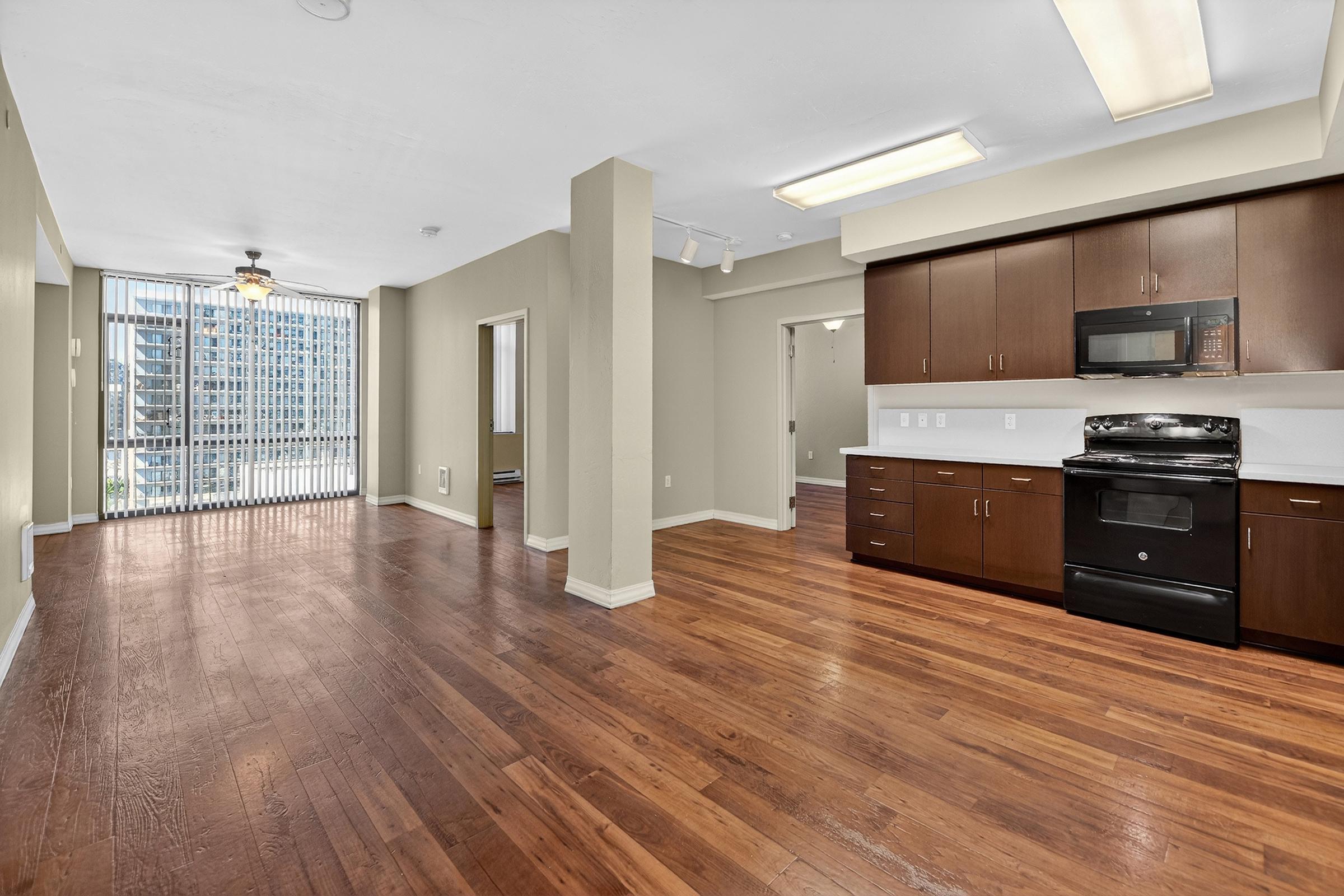 Bright, spacious apartment interior featuring hardwood floors, modern kitchen with dark wood cabinets, black appliances, and natural light from large windows. The open layout includes a living area and transition to the kitchen, creating a welcoming atmosphere.