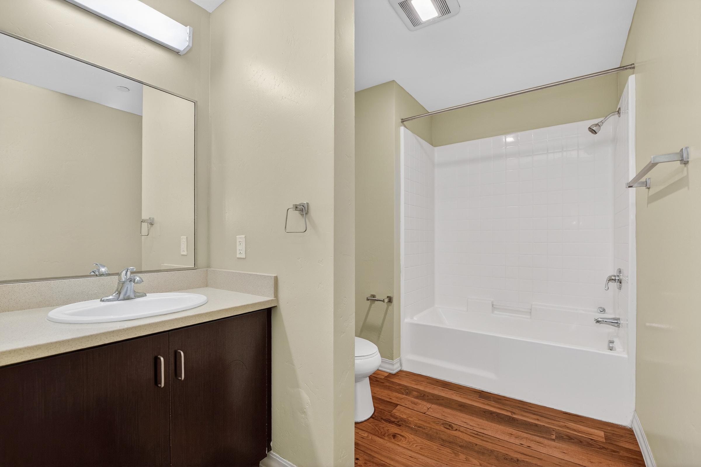 A modern bathroom featuring a single sink vanity with a mirror above, and a separate area with a bathtub and shower. The walls are painted a light beige, and the floor has wooden planks. Essentials like a toilet and towel bar are visible.