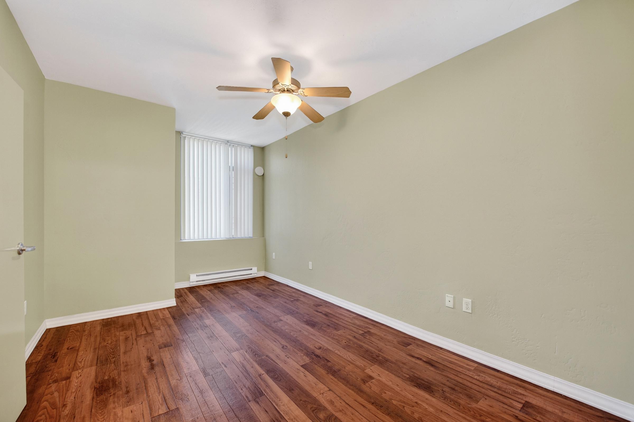 A well-lit, empty room featuring light green walls, hardwood flooring, and a ceiling fan. A large window with vertical blinds allows natural light to enter. The space appears clean and ready for furnishing, with a simple, minimalist design.
