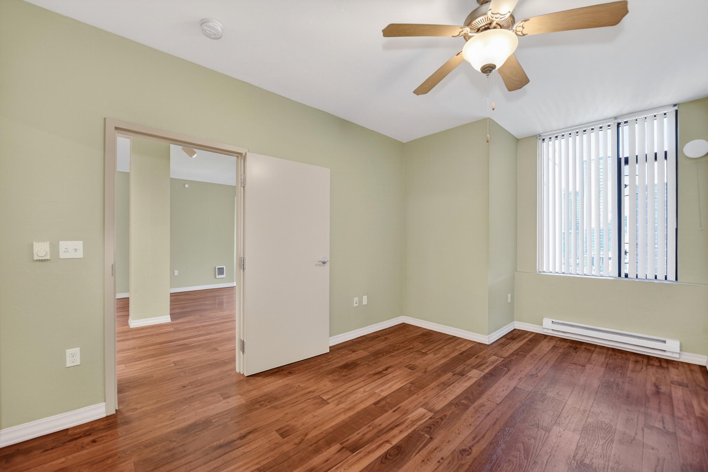 A spacious, well-lit room with green walls and hardwood flooring. It features a ceiling fan, an open doorway leading to another area, and large windows with vertical blinds. The layout suggests a modern design, with bright natural light streaming in.