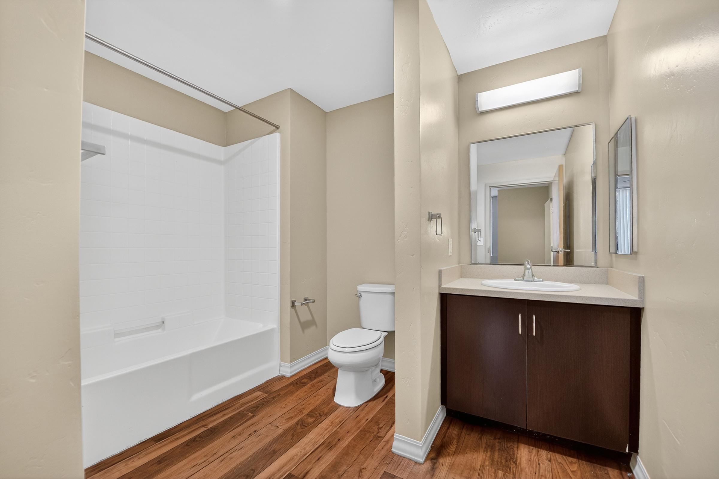 Modern bathroom featuring a shower-tub combination, a toilet, and a vanity with a sink. The walls are painted a light beige, and there is a large mirror above the sink. Wooden flooring adds warmth to the space, and a soft light fixture illuminates the area.