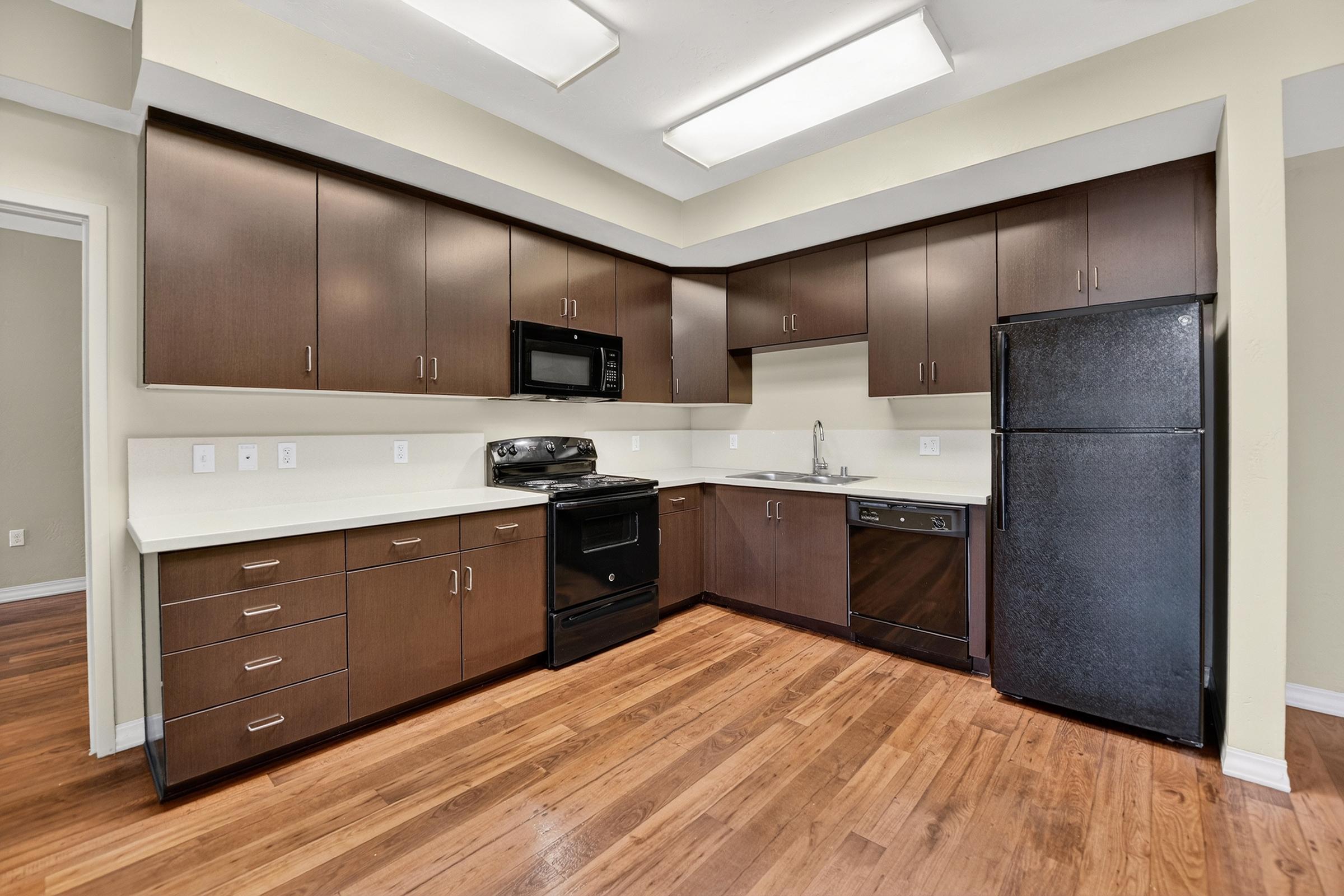 Modern kitchen with dark wood cabinetry, white countertops, and stainless steel appliances. Features a black refrigerator, microwave, oven, and dishwasher. The floor is hardwood, and the walls are painted a light beige. Ample cabinet space and a minimalistic design create an open, inviting atmosphere.