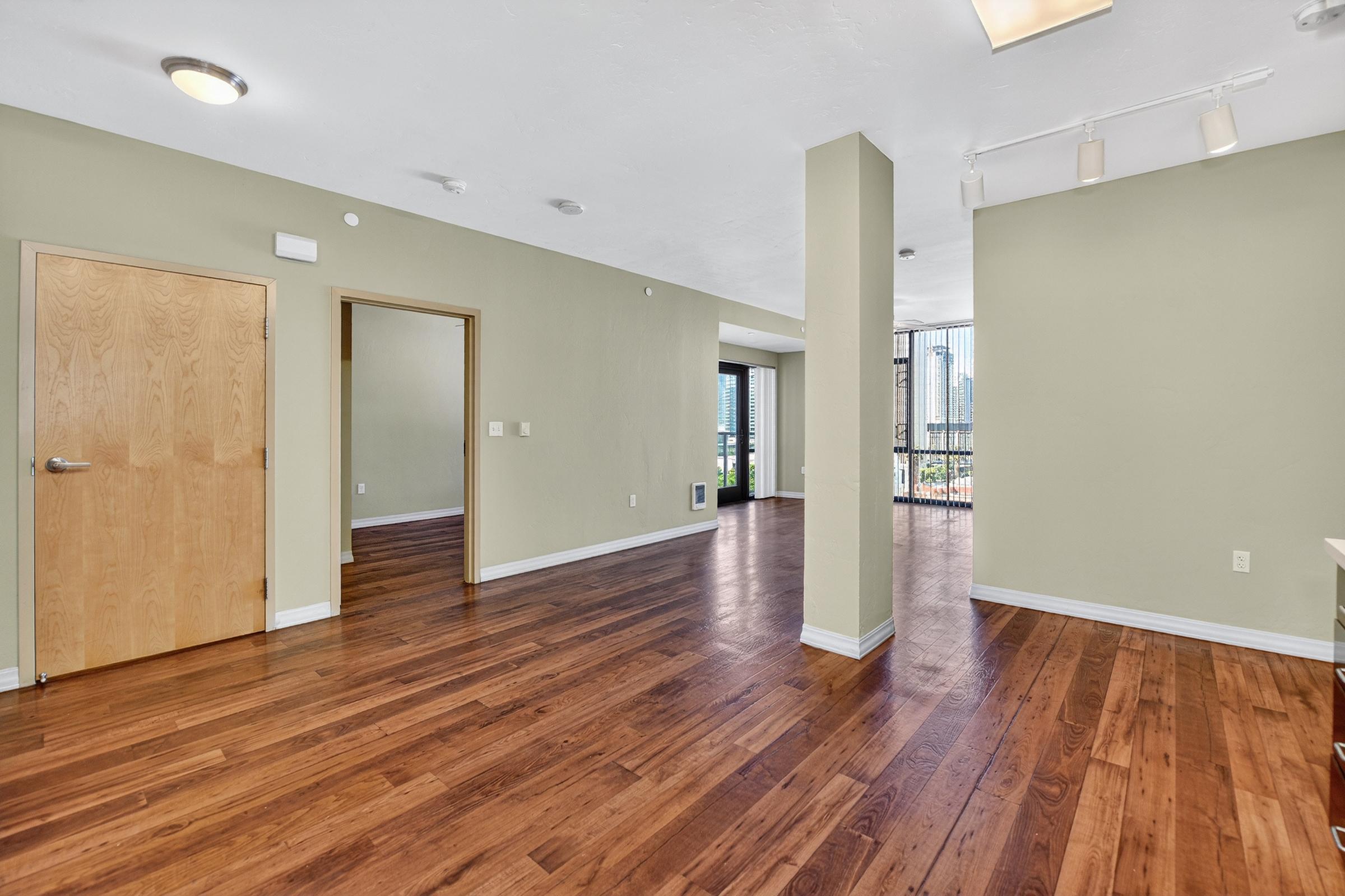 A spacious, empty interior room with wooden flooring. There are two doorways visible, one leading to a hallway and the other to a balcony. Natural light streams through large windows, highlighting the light green walls and modern lighting fixtures above. The room appears ready for furniture placement.