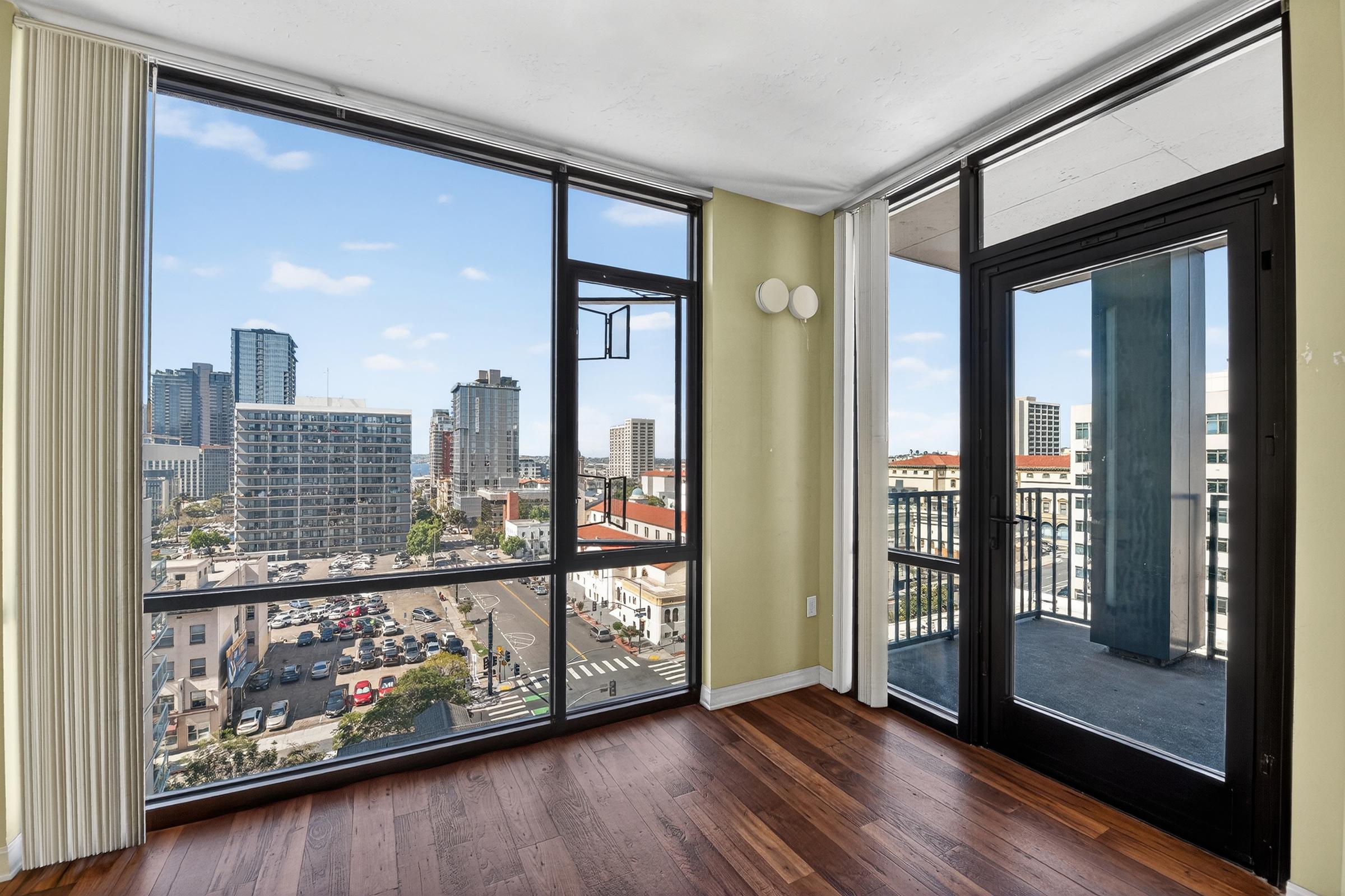 Interior view of a modern room with large windows showcasing a city skyline. Natural light fills the space, highlighting hardwood floors and minimalistic decor. The balcony door leads to an outdoor area, while buildings and streets are visible in the background under a clear blue sky.