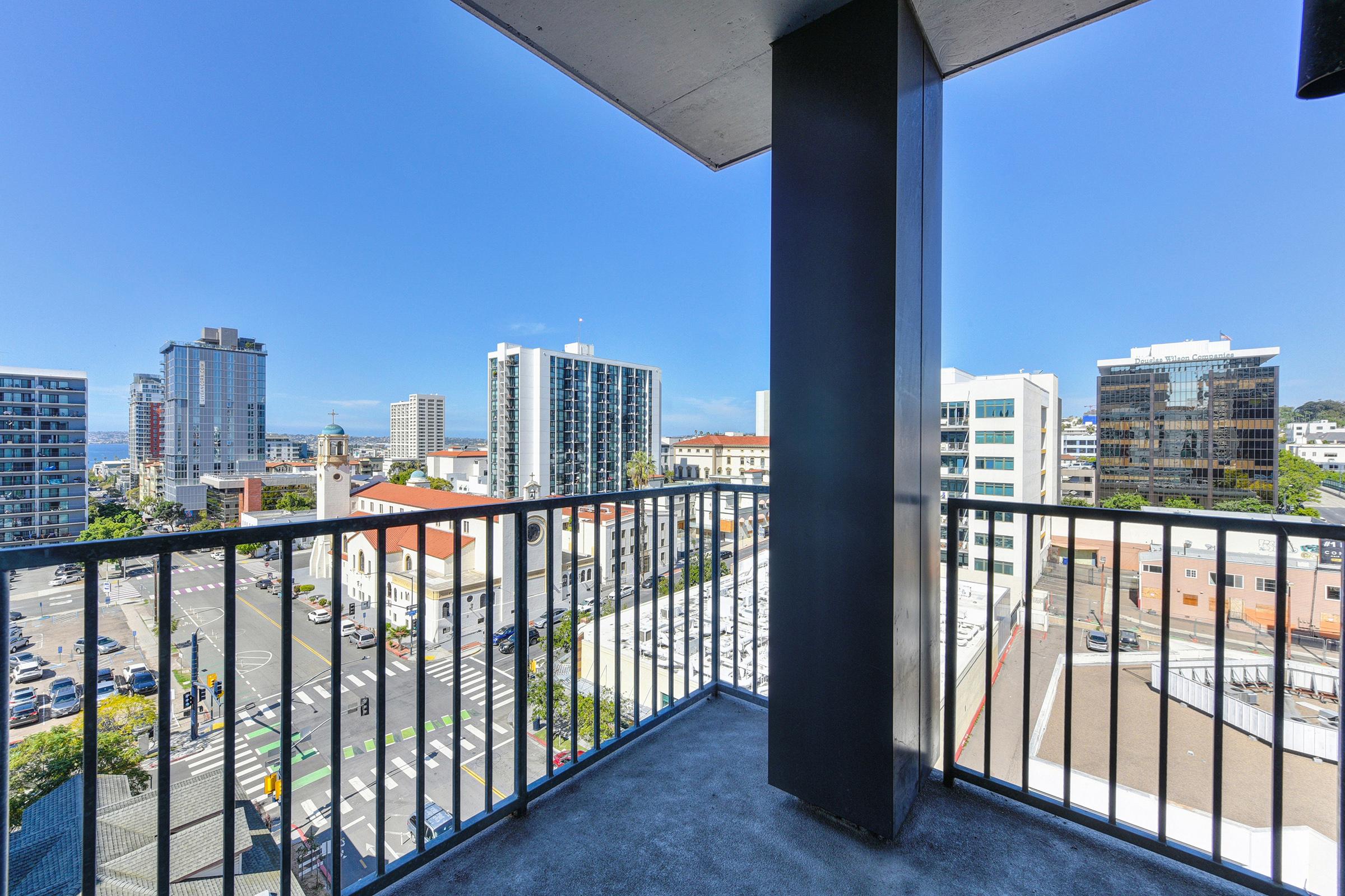 View from a balcony overlooking a city skyline. The scene includes tall buildings, a clear blue sky, and a bustling street below with cars and pedestrians. The foreground features a metal railing, emphasizing the perspective from the balcony.