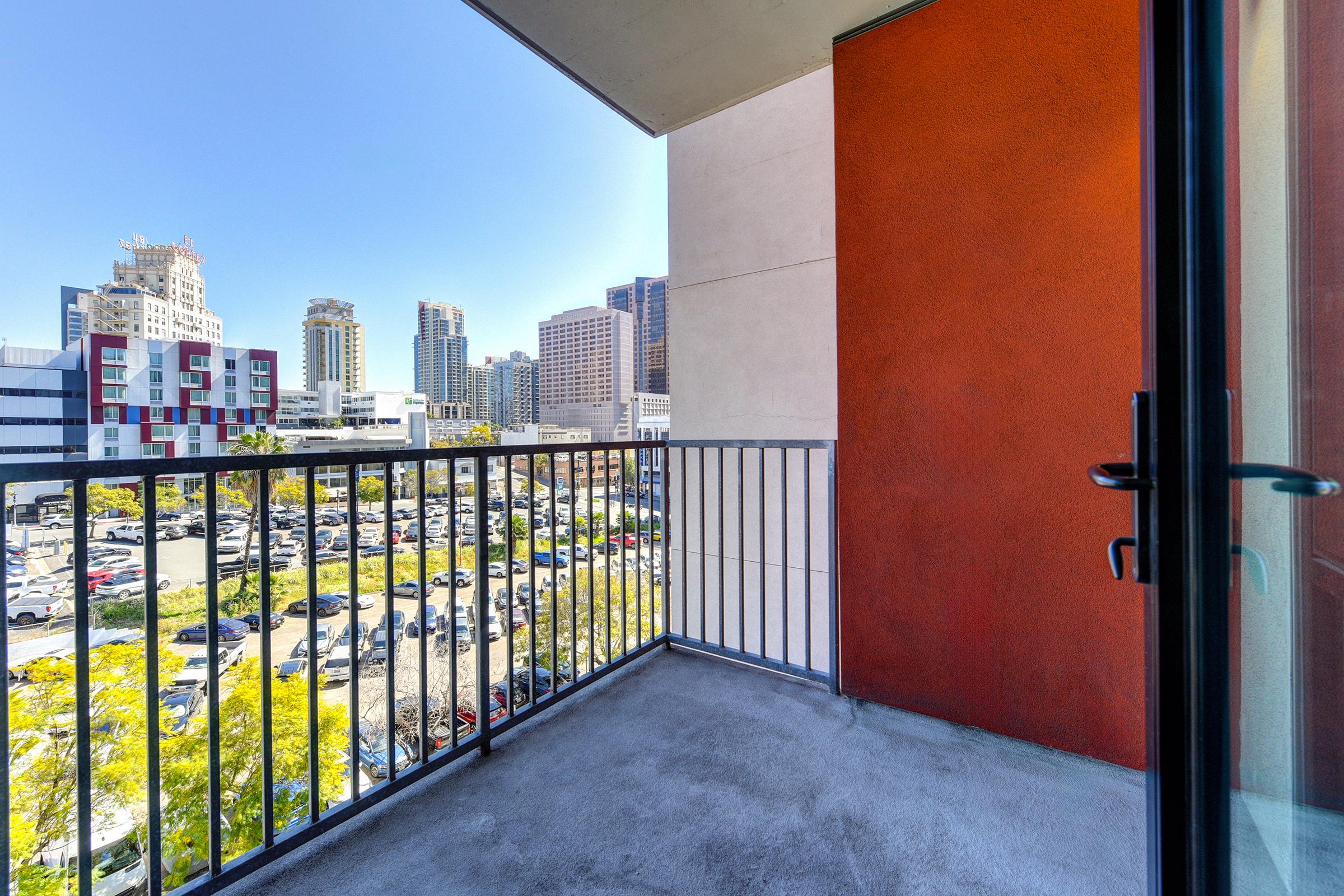 A modern balcony view overlooking a parking lot and city skyline. The balcony features metal railings and is adjacent to a vibrant orange wall. Skyscrapers and greenery are visible in the background under a clear blue sky.