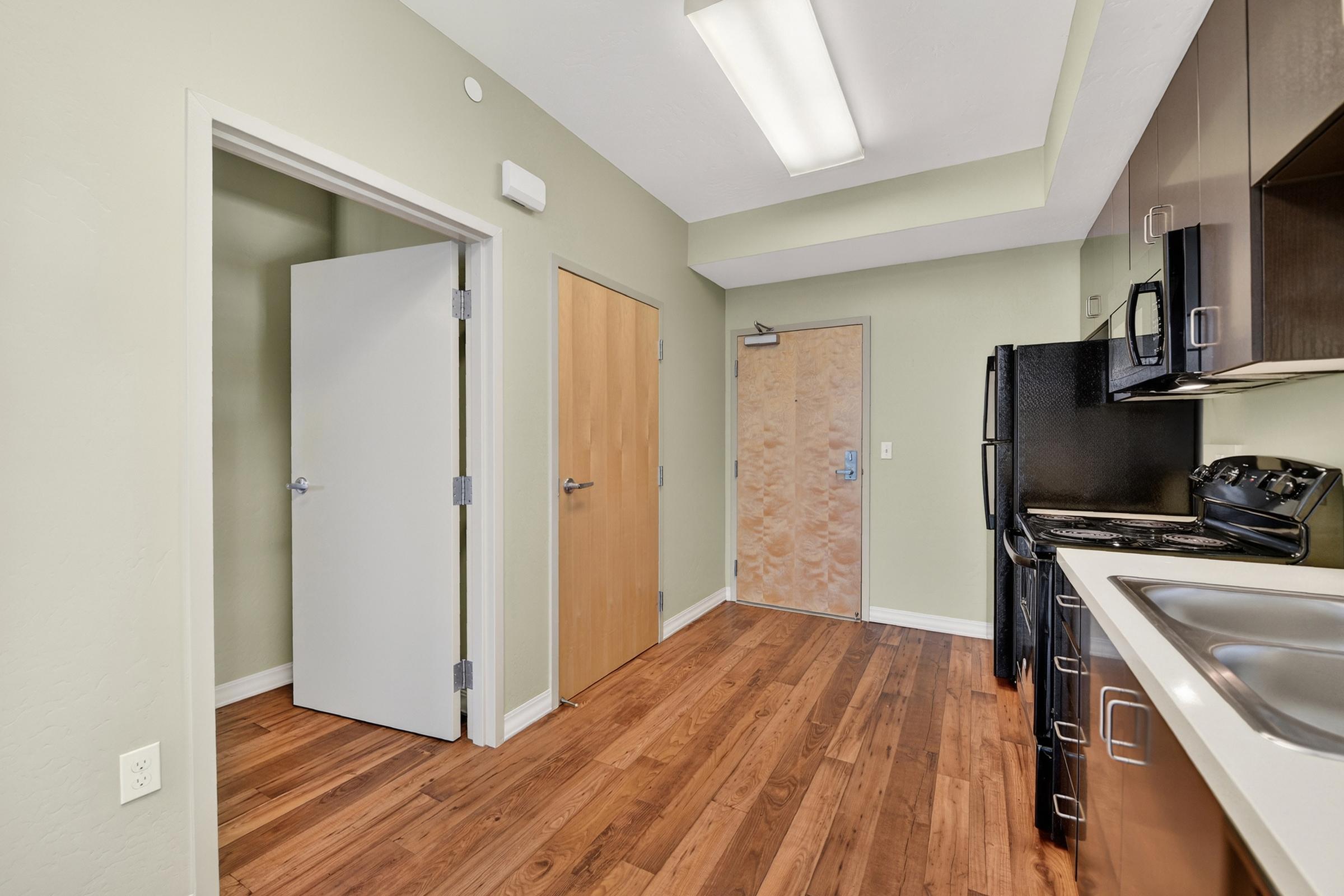 Modern kitchen and entryway with wood flooring. The kitchen features dark cabinets, a stainless steel sink, and a stove. A closed door leads to a room, while another door opens to the hallway. Natural light from overhead lights illuminates the space, creating a bright and inviting atmosphere.