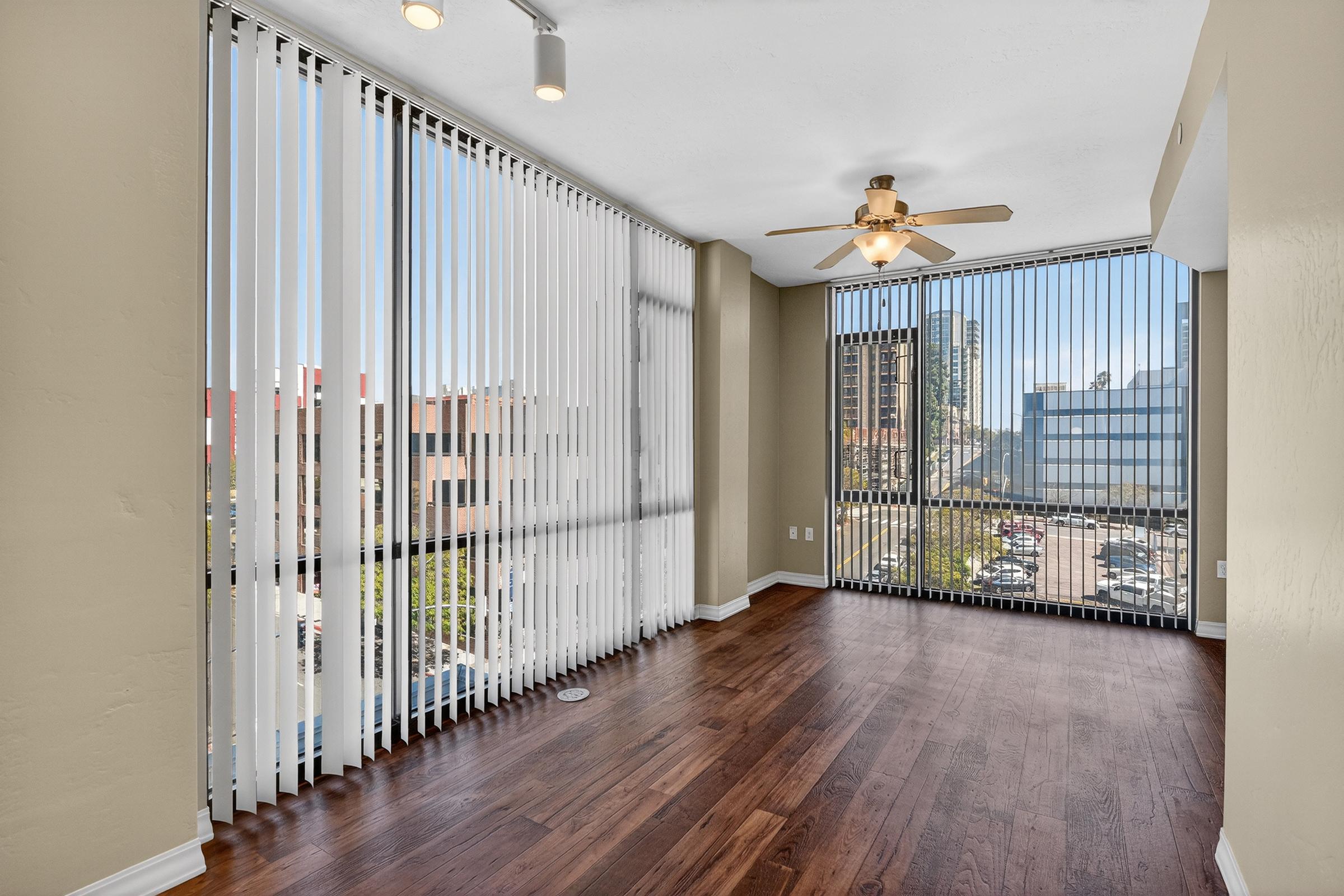 Bright, spacious room with wooden flooring and large vertical blinds allowing natural light. A ceiling fan hangs from the ceiling, and the view outside features a cityscape. The walls are painted in a neutral color, creating a warm and inviting atmosphere.