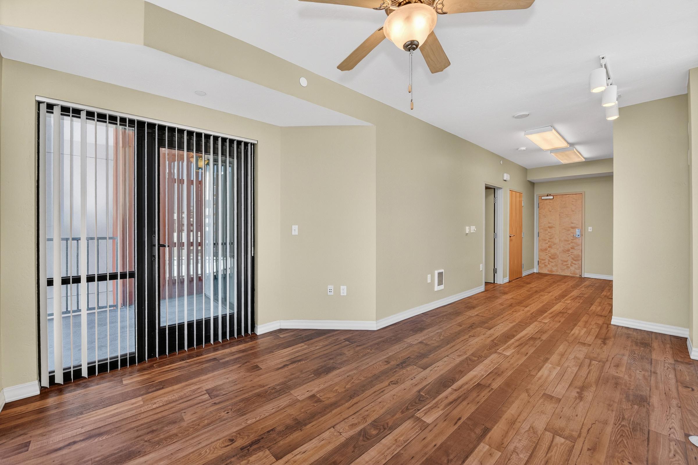 Interior view of a spacious room featuring wooden flooring, light beige walls, and a ceiling fan. To the left, there are large sliding doors with vertical blinds leading to a balcony. The room is well-lit and includes doorways leading to other areas, such as a bathroom or additional rooms.