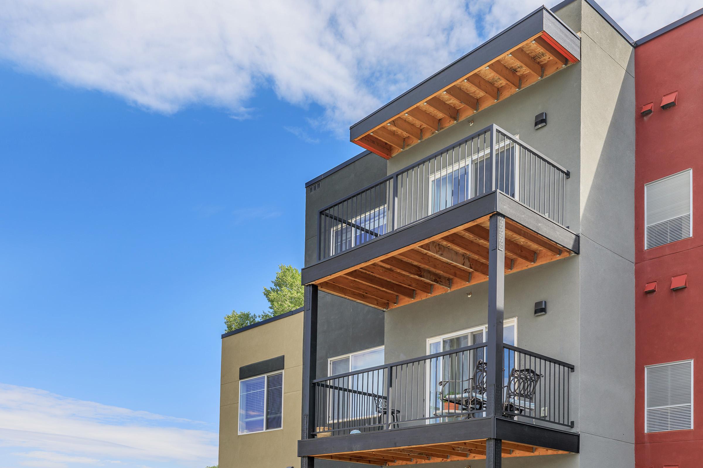 A modern multi-story building with a sleek design, featuring balconies on the second and third floors. The upper balcony has a wooden ceiling, and the exterior includes contrasting colors, with one side in gray and the other in red. The sky is clear and blue, enhancing the contemporary look of the structure.